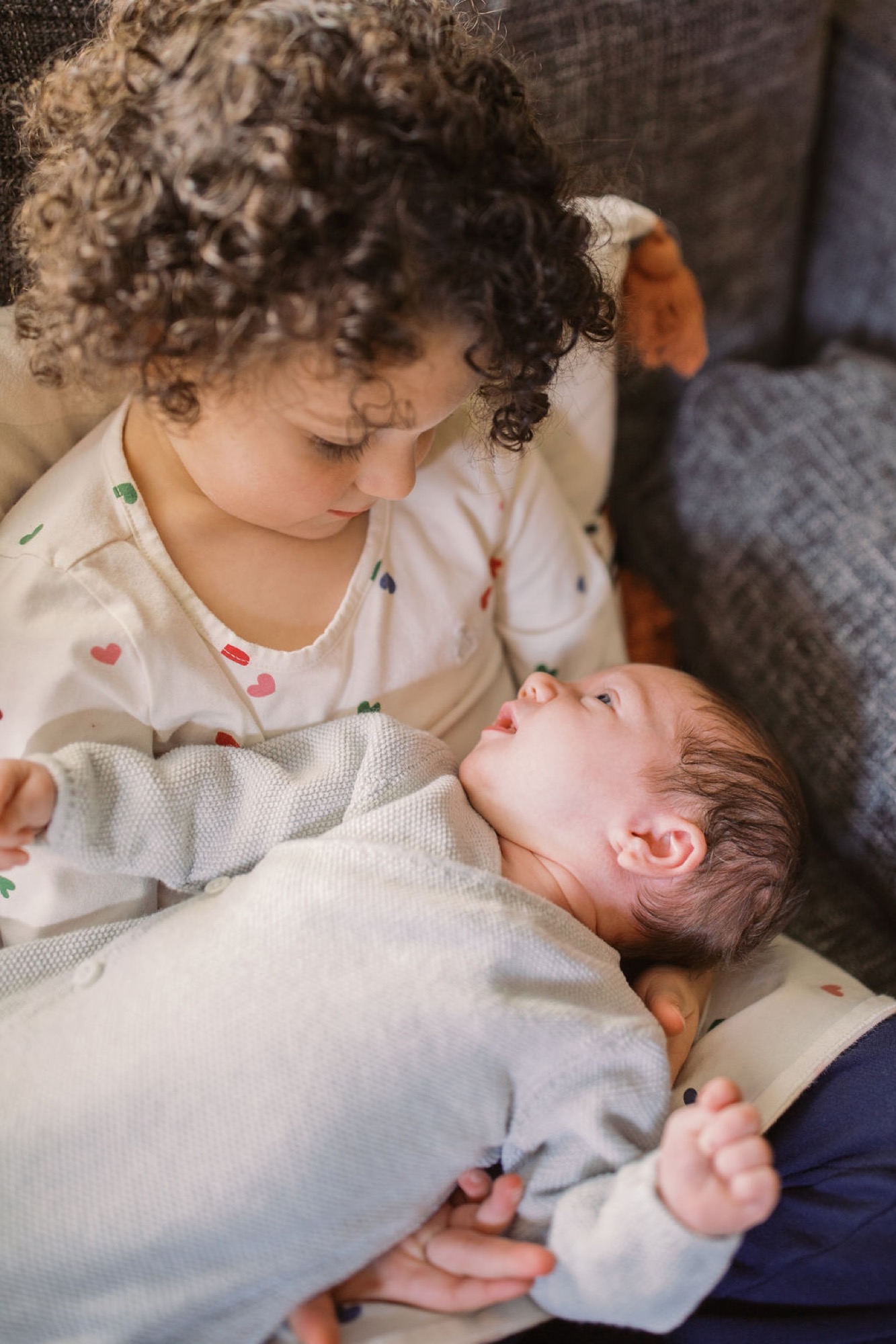 Older sister gazes tenderly at newborn brother in her arms, wearing matching patterned shirts in soft indoor light