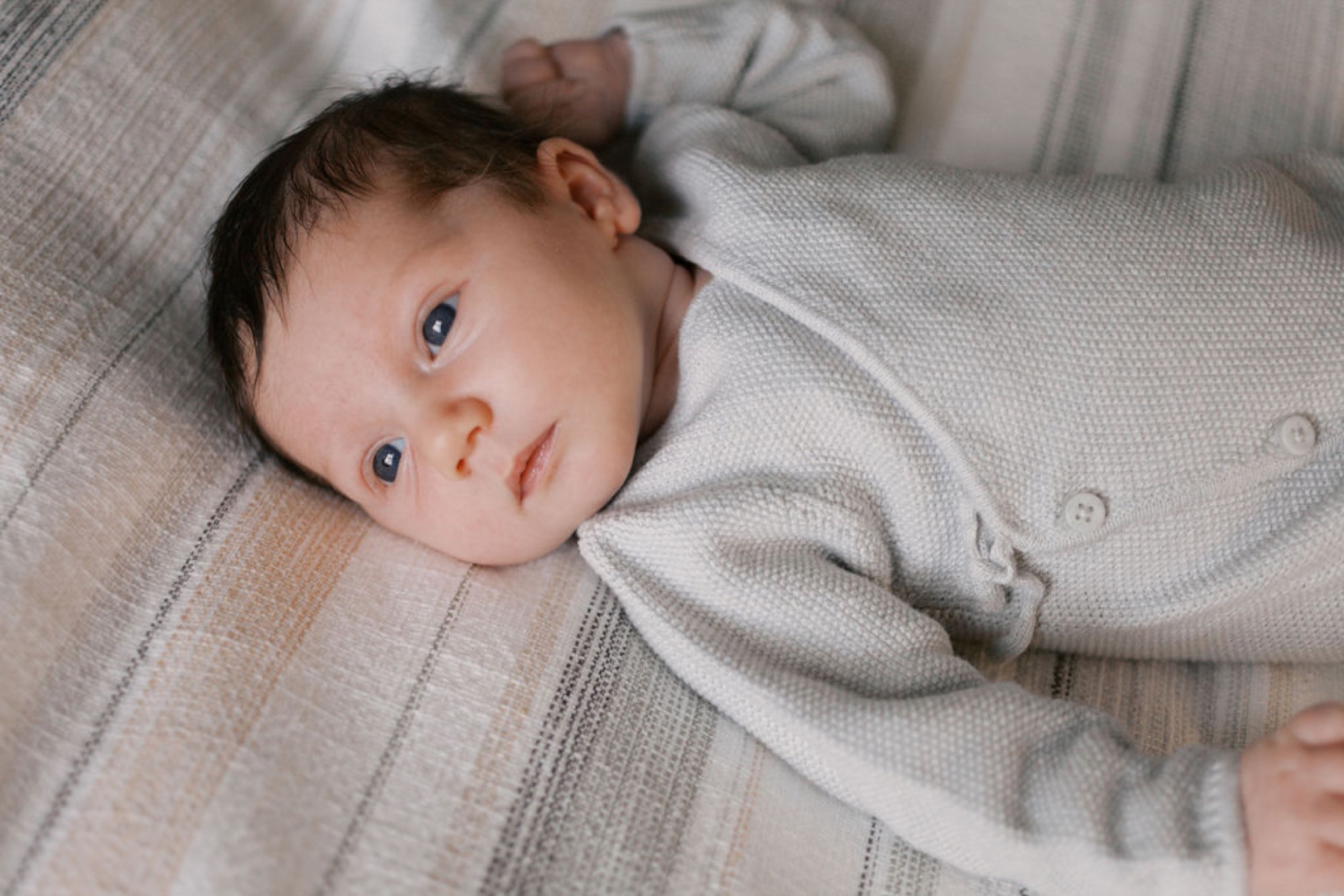 Newborn with dark eyes gazes upward while swaddled in soft gray knit, lying on neutral striped bedding in warm natural light