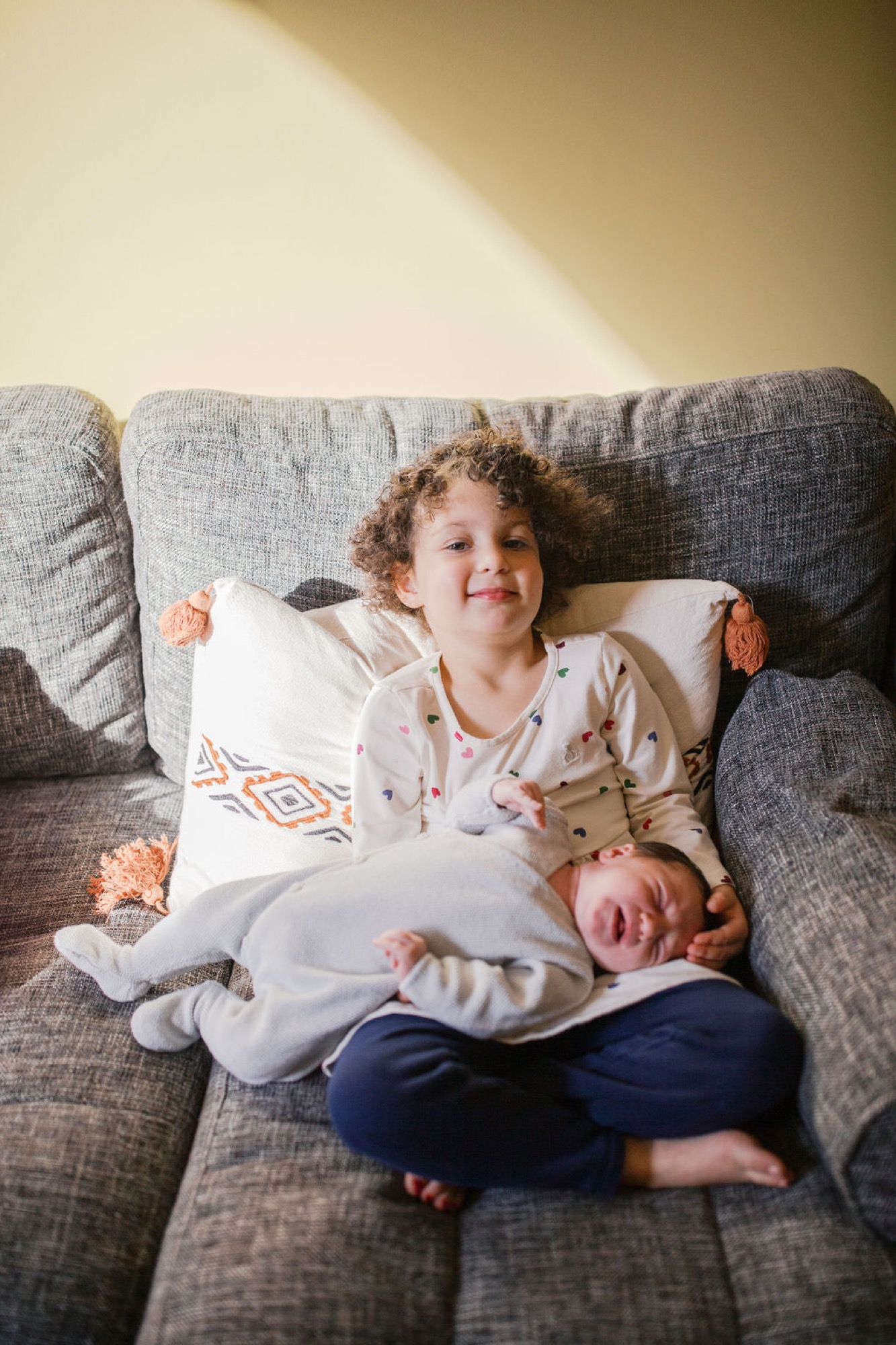 Toddler with curly hair beams at camera while cradling sleeping newborn on a gray linen couch bathed in warm sunlight