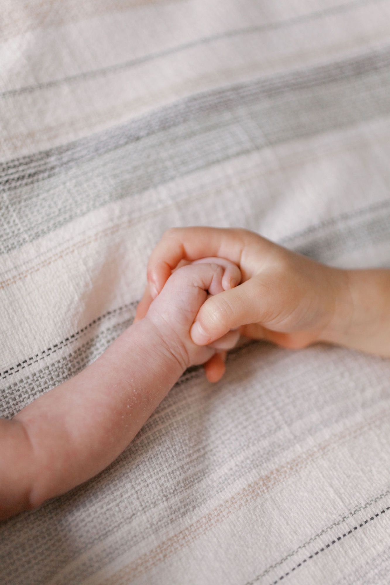Tiny newborn hand grasps parent's finger against soft striped linen, capturing tender first moments of connection