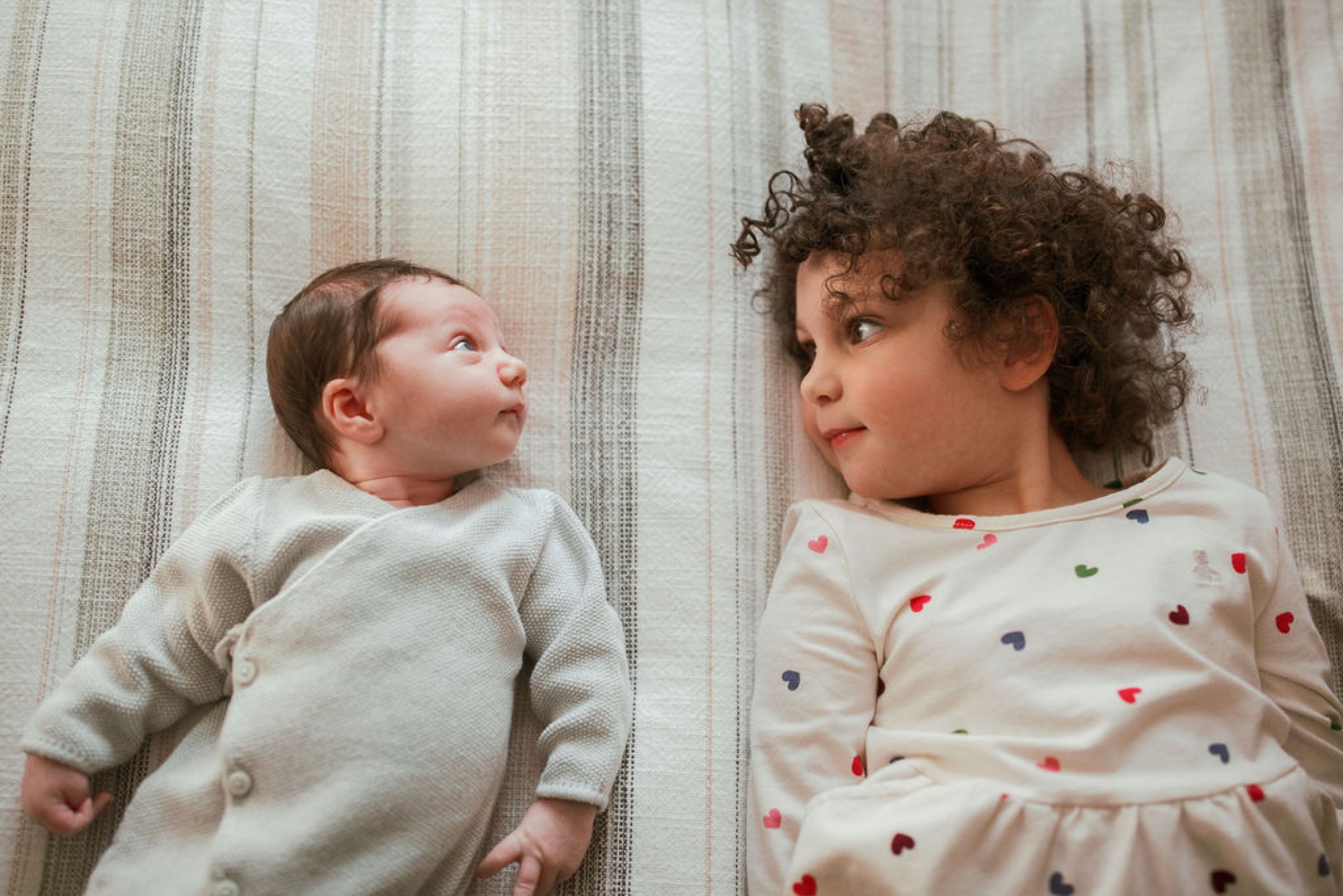 Two young children lie on a striped neutral fabric, gazing upward with thoughtful expressions while wearing coordinating cream-colored clothing