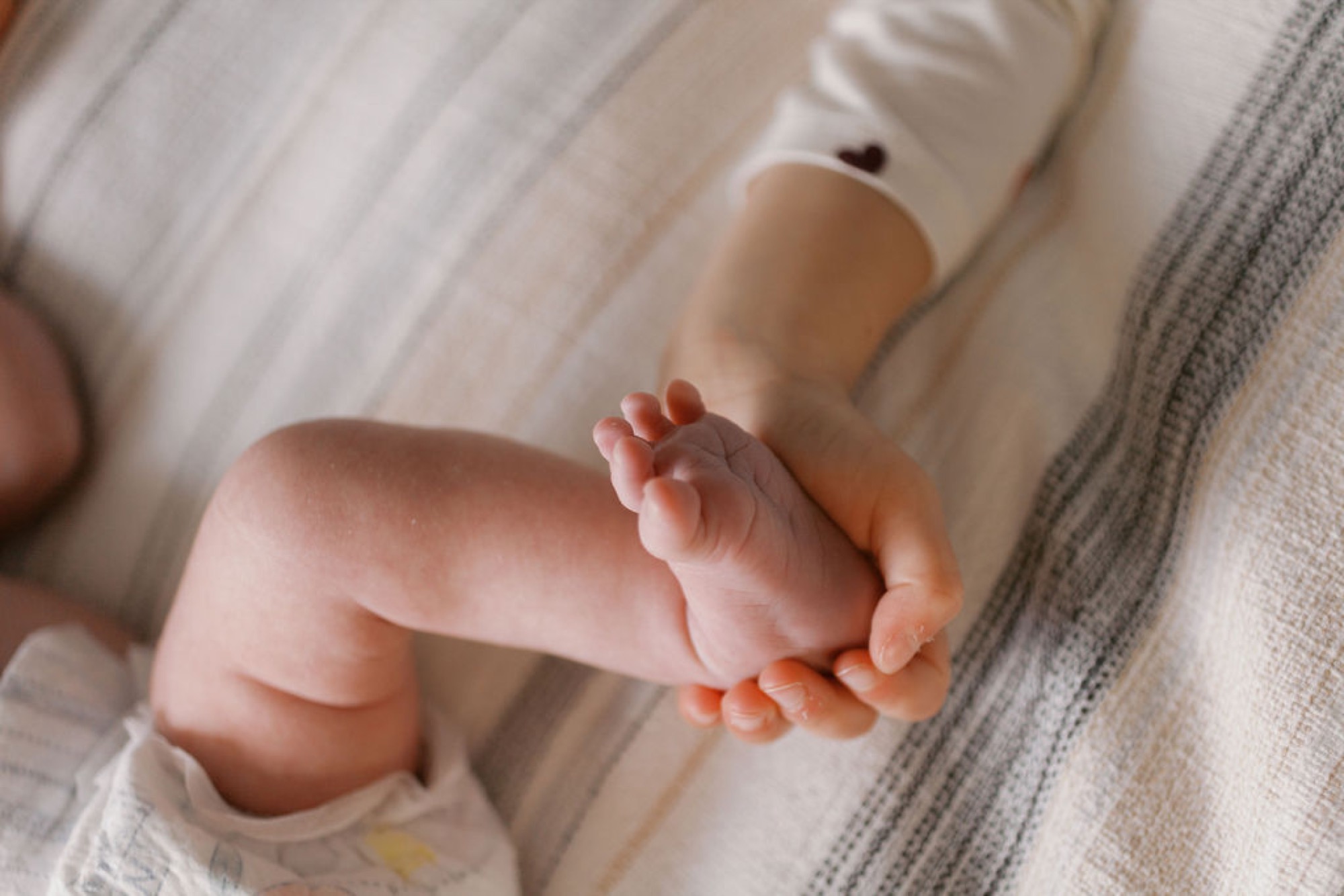A newborn's delicate foot rests in an adult's open palm, soft natural light illuminating the tiny toes against neutral bedding