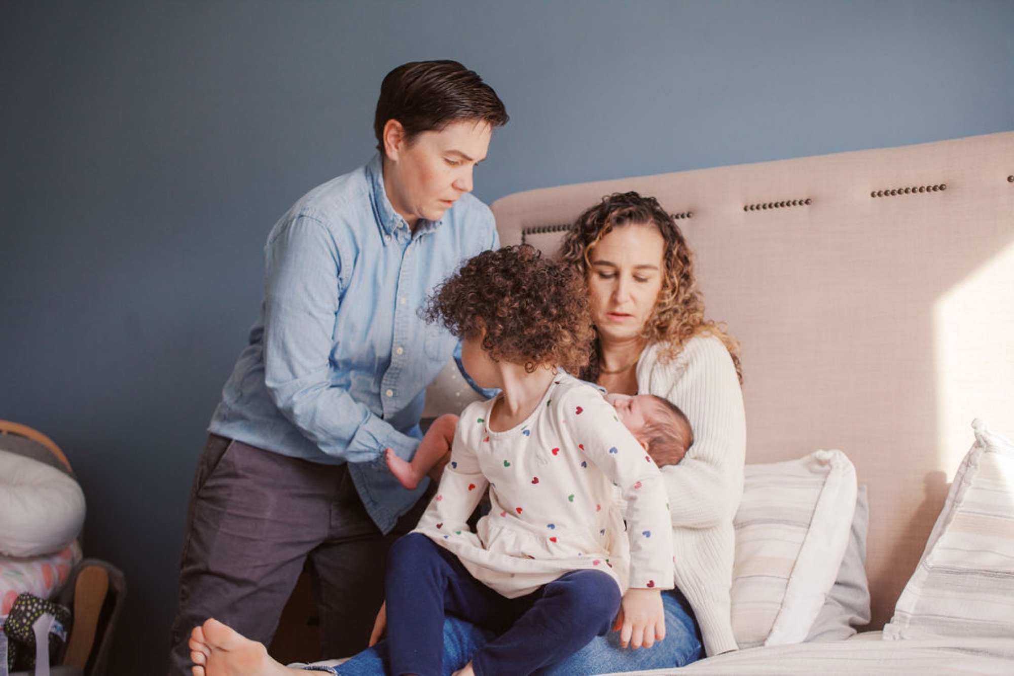 One mom gazes at newborn while the other holds the infant, older daughter sits between them on cream upholstered bed with soft natural light