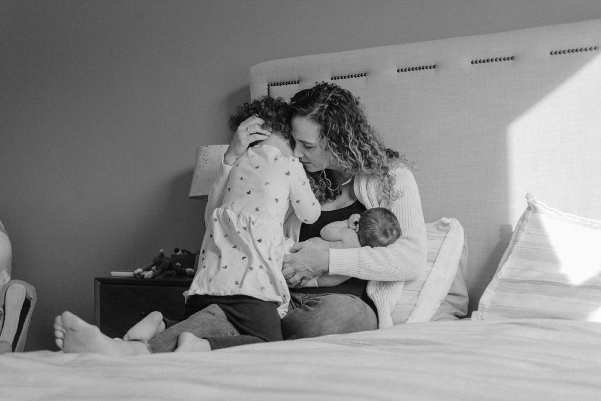A mother sits on a bed holding her newborn while her older child leans in close, kissing the baby's head in tender affection
