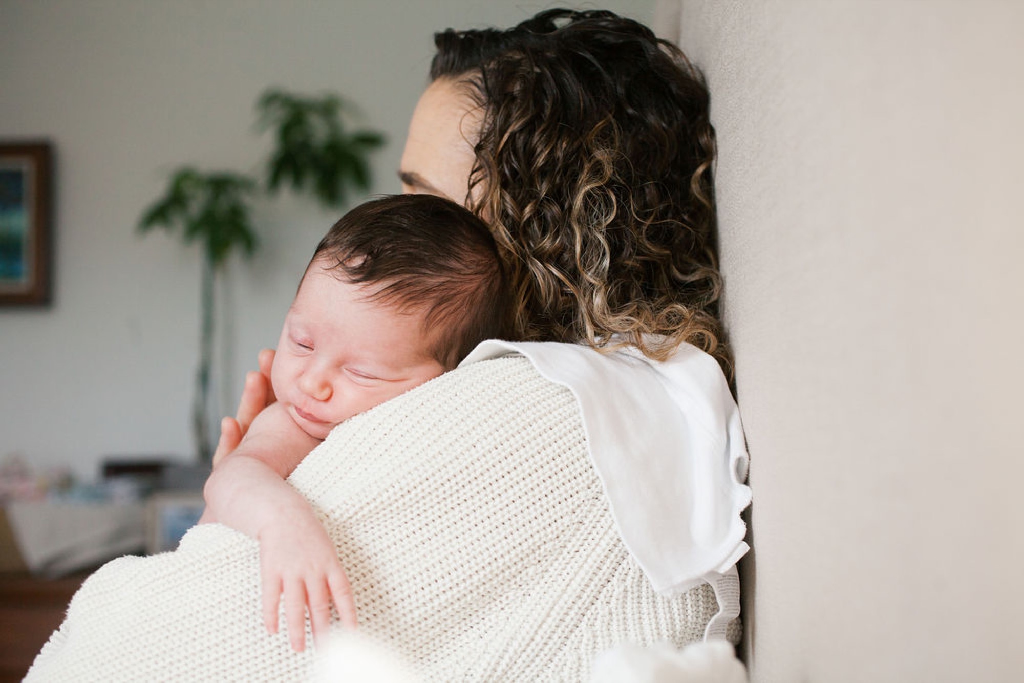 A newborn sleeps peacefully against a parent's shoulder in a cream knit sweater, captured in soft natural light from an indoor session