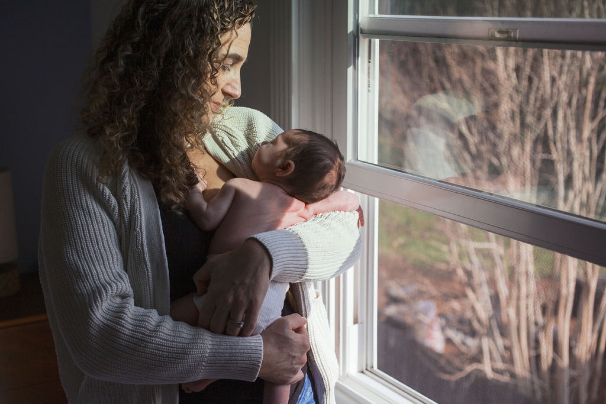 Mother gazes down at newborn wrapped in cream knit while soft window light illuminates their profile against rolling hillside landscape