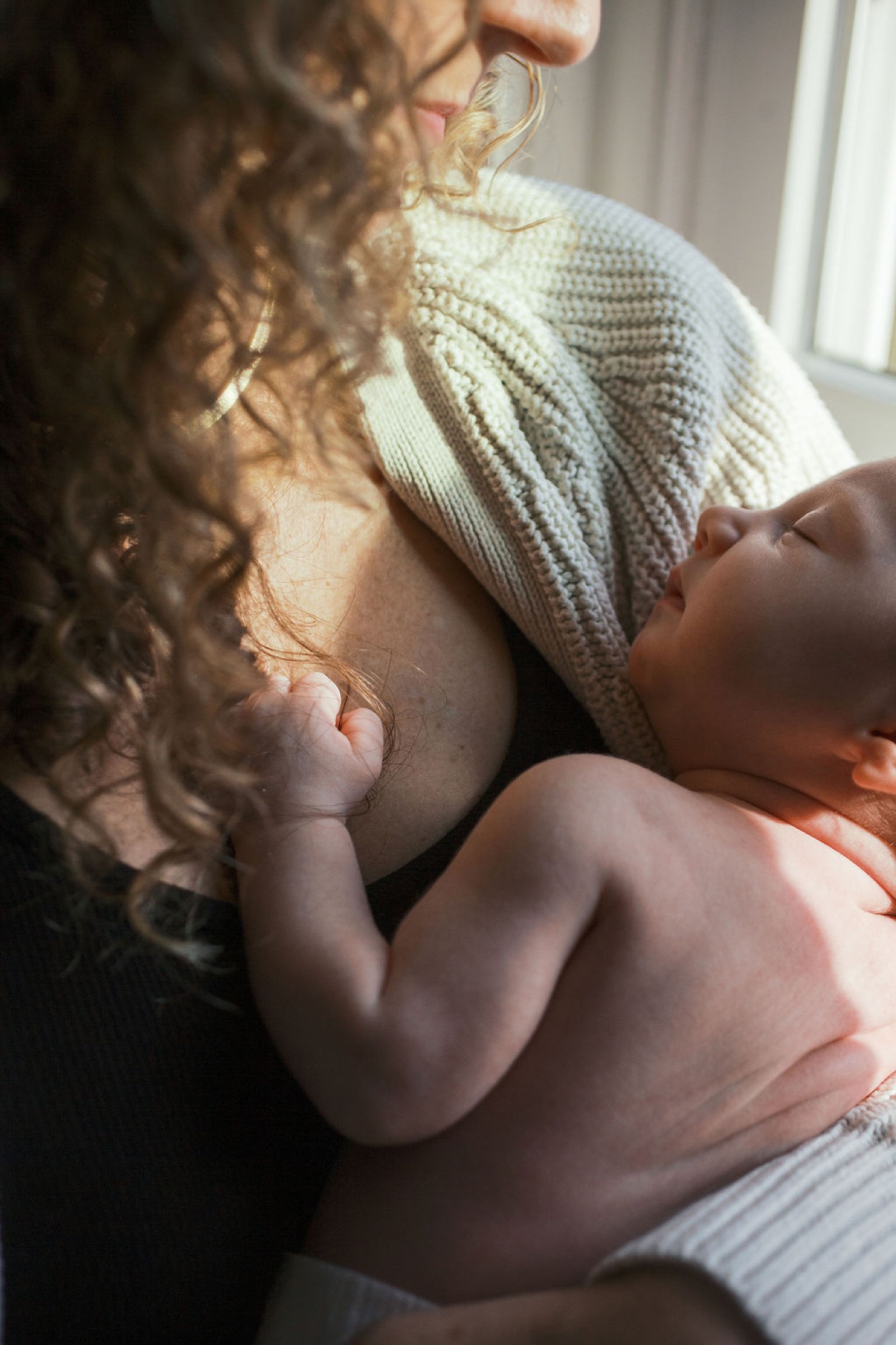 A newborn's tiny hand grasps her mother's cream-knit sweater while nursing in warm natural window light