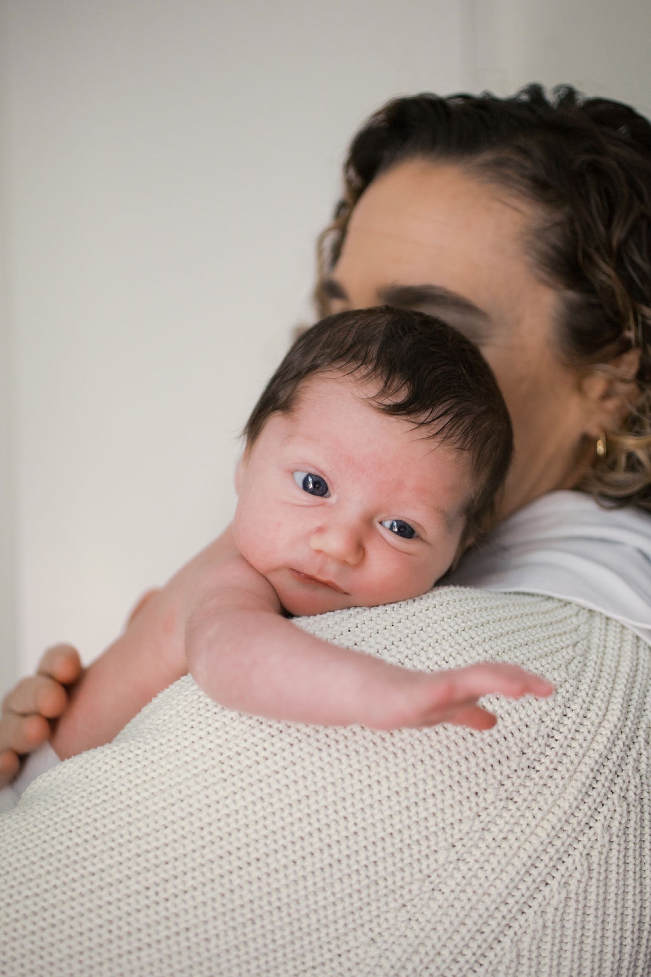 Newborn with alert, wide eyes gazes at camera while cradled against mother's cream knit shoulder in soft natural light