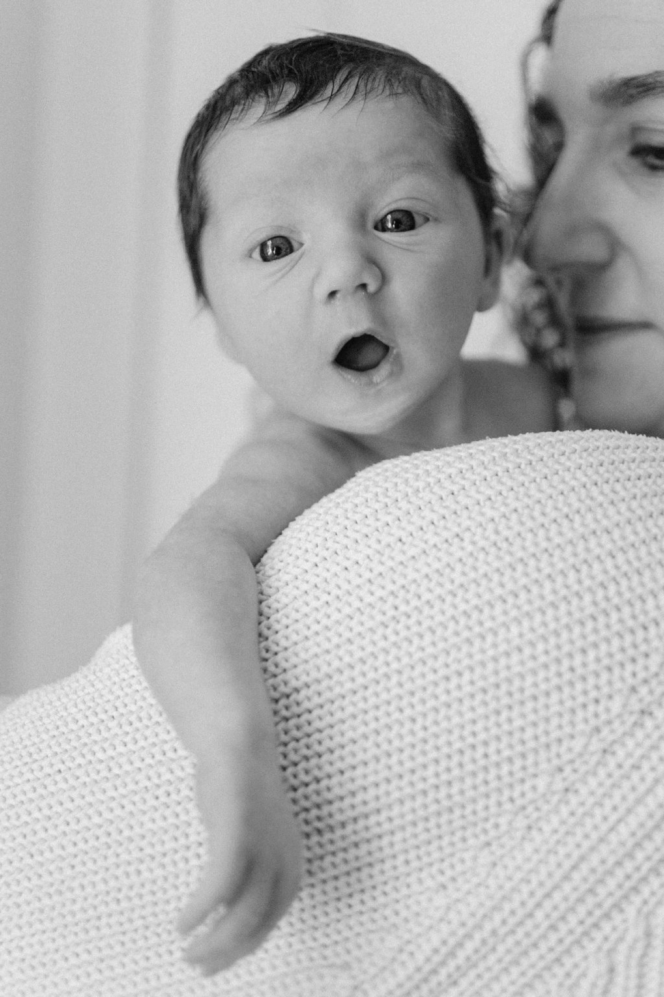 Newborn gazes wide-eyed over parent's shoulder, mouth open in wonder, wrapped in soft cream blanket during intimate indoor session
