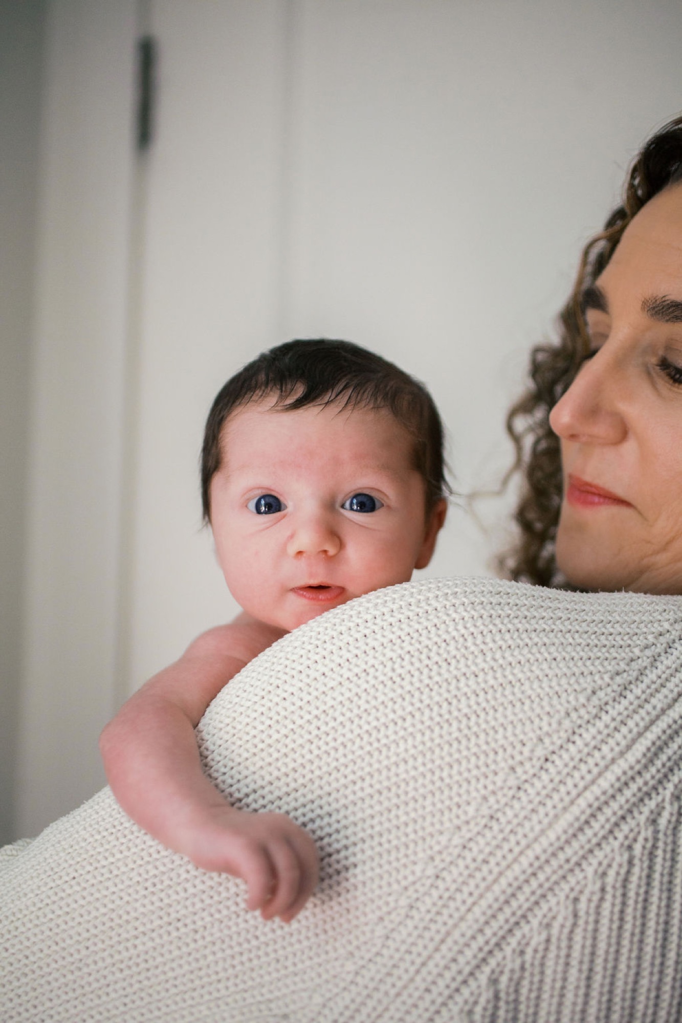 Newborn with wide alert eyes gazes toward camera while cradled against mother's cream knit shoulder in soft natural light
