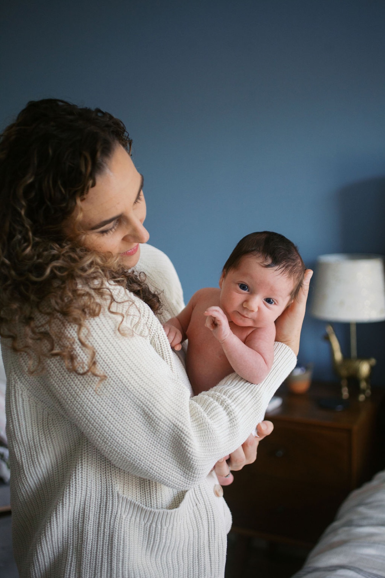 A mom in cream cardigan holds newborn against her shoulder, baby gazing at camera with alert expression against deep blue bedroom wall