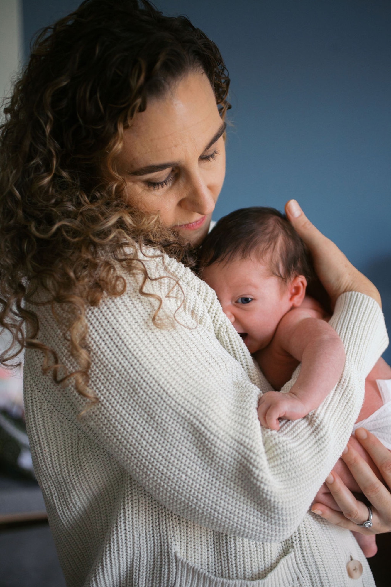 Mother in cream knit cardigan gazes down tenderly at newborn cradled against her shoulder against soft blue backdrop