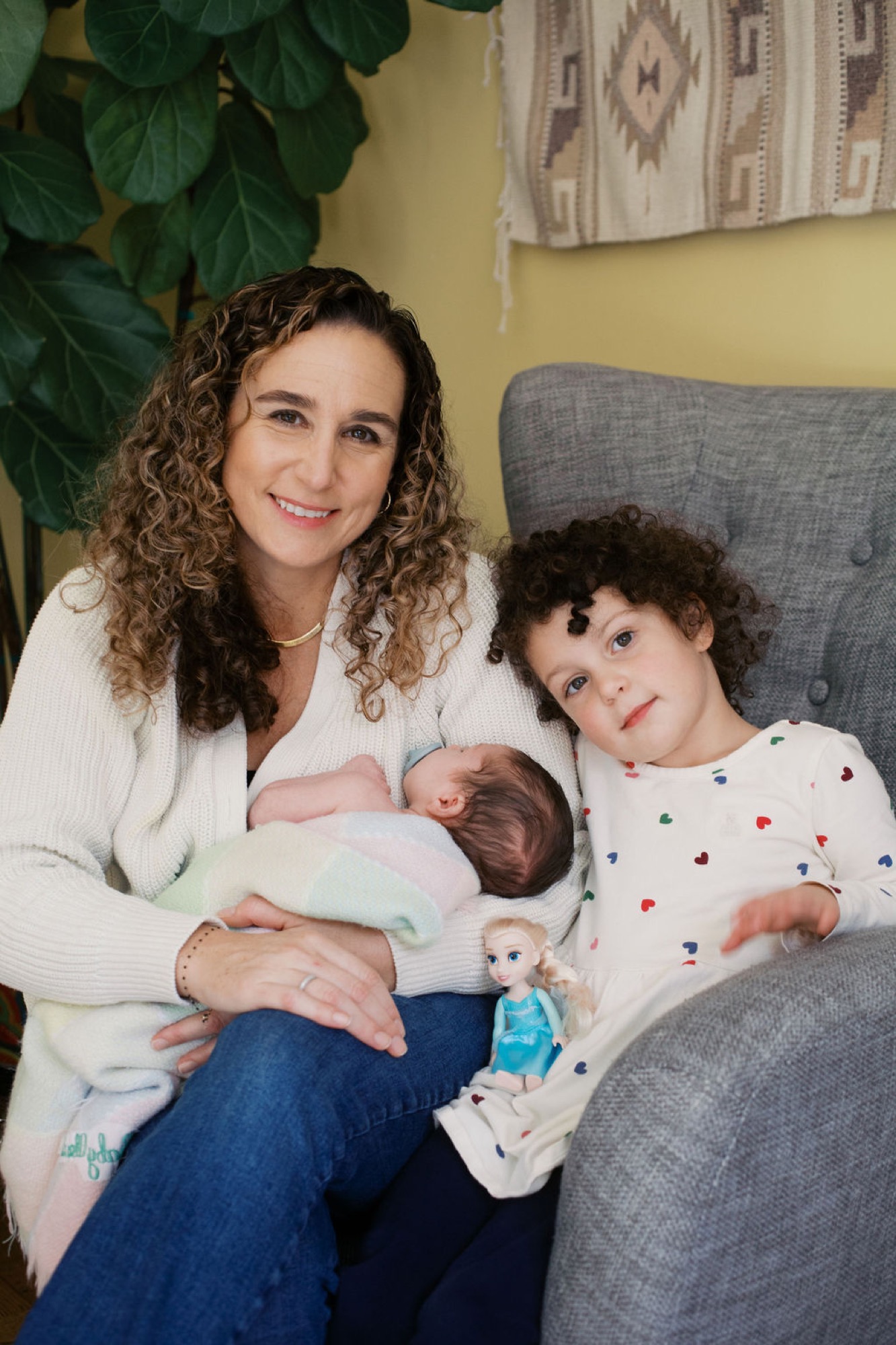 Mother smiles warmly while cradling newborn in cream cardigan, older child nestled beside them on gray chair against yellow wall
