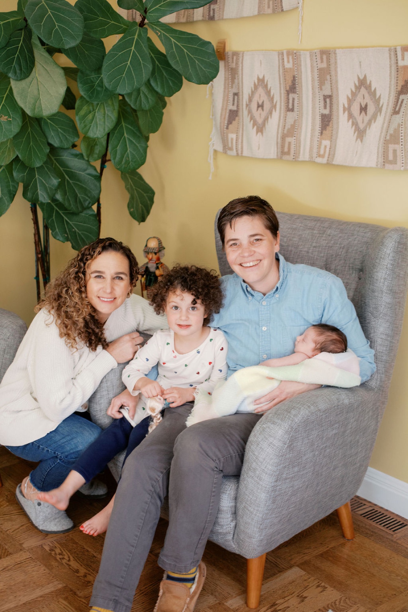 Family of four smiles warmly on a gray chair against a yellow wall, with dad cradling newborn while mom and toddler sit close beside them