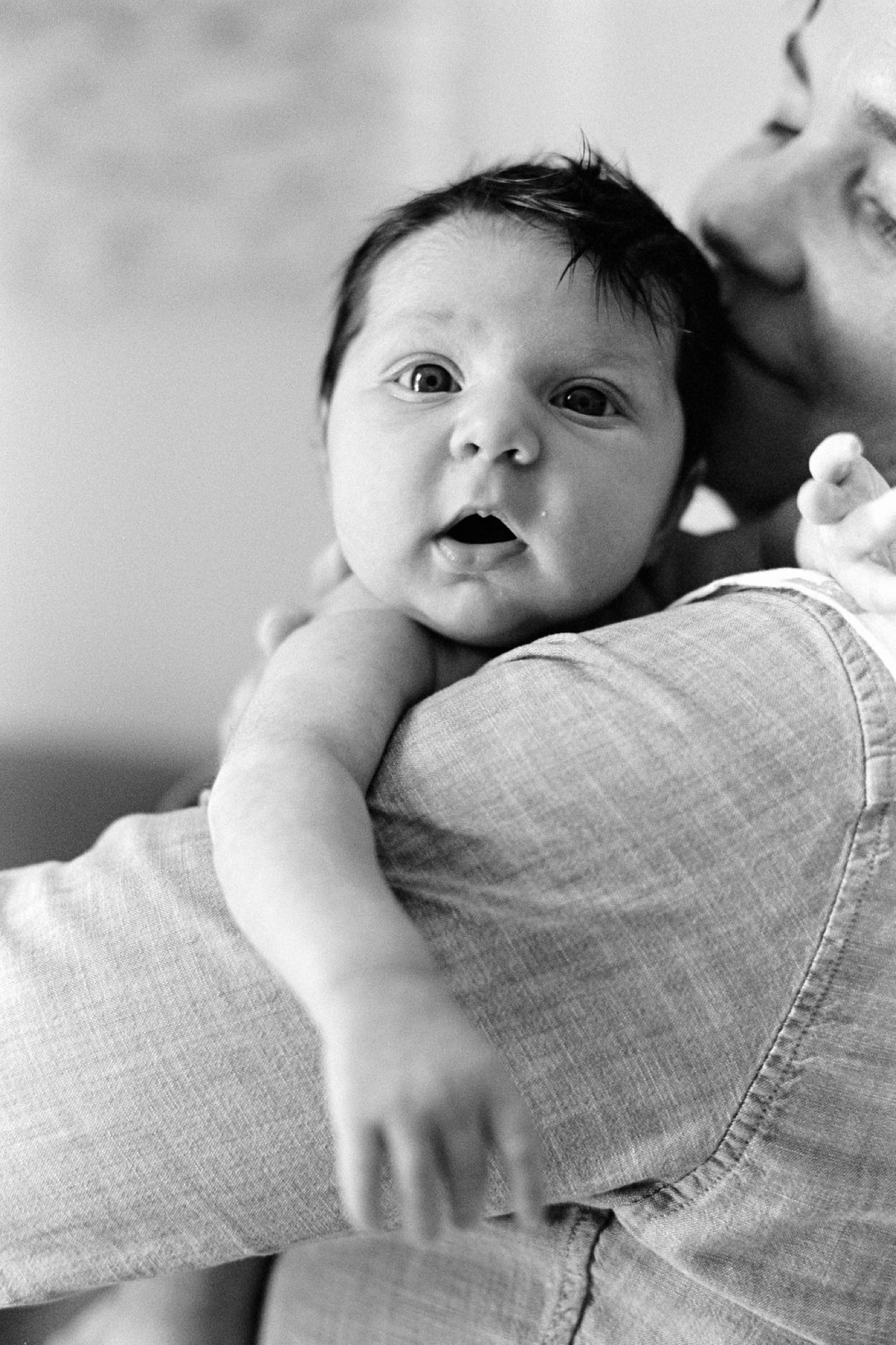 Newborn gazes upward with wide-eyed wonder, cradled against parent's shoulder in soft natural light