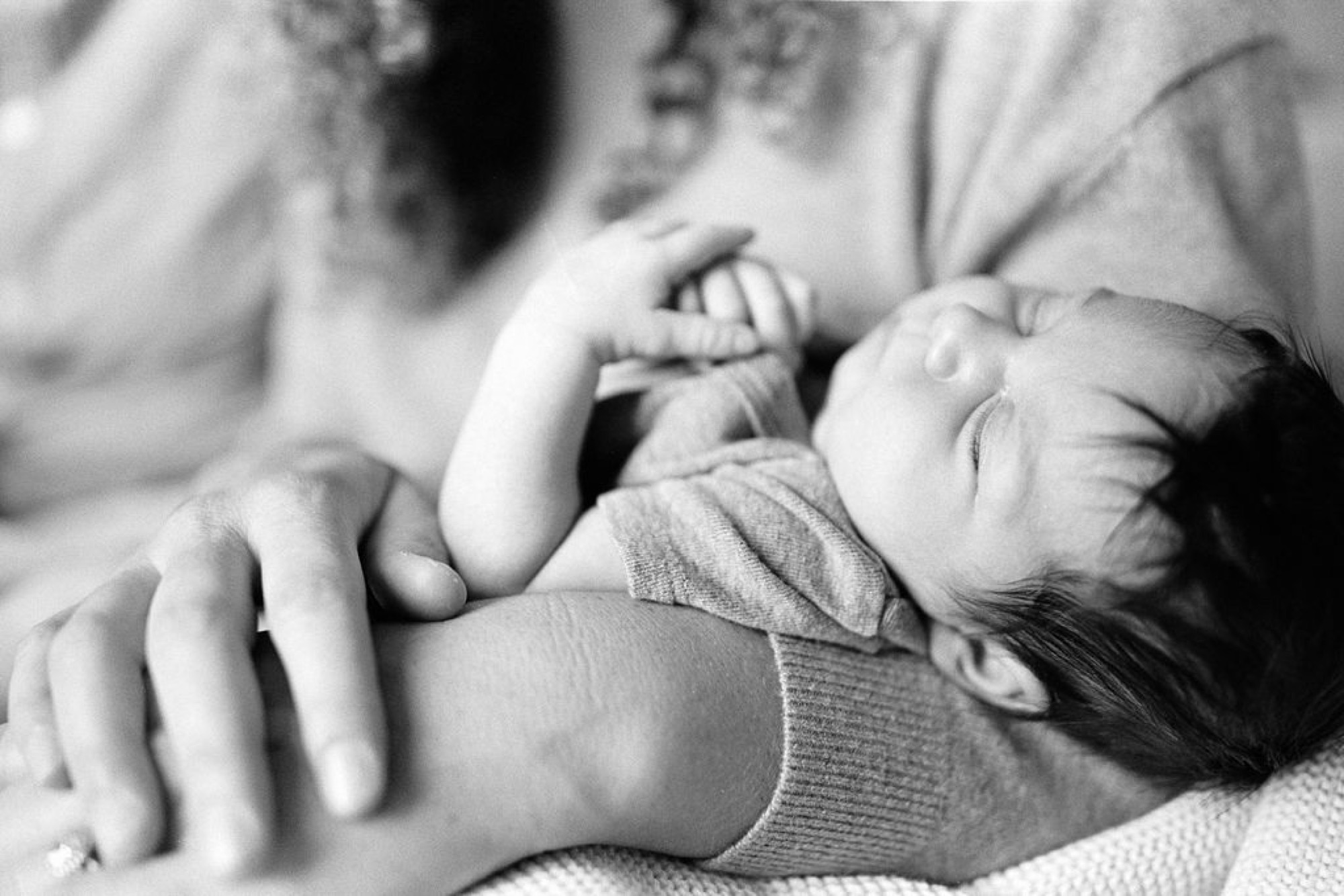 Newborn's tiny hand grasps parent's finger in tender black-and-white portrait, soft natural light illuminating delicate skin and peaceful sleeping expression