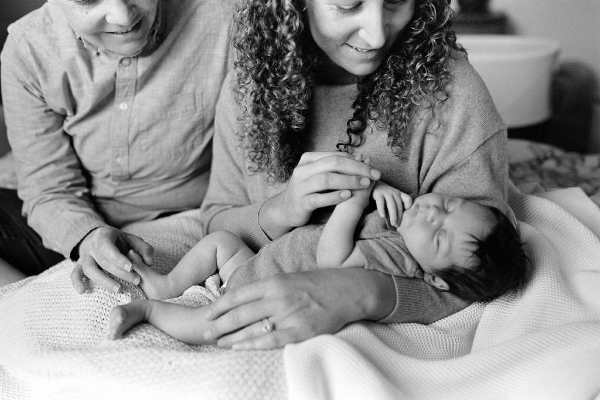 Two sisters with curly hair gently touch their newborn brother's hand, their genuine smiles reflecting pure joy as he sleeps peacefully on a soft blanket