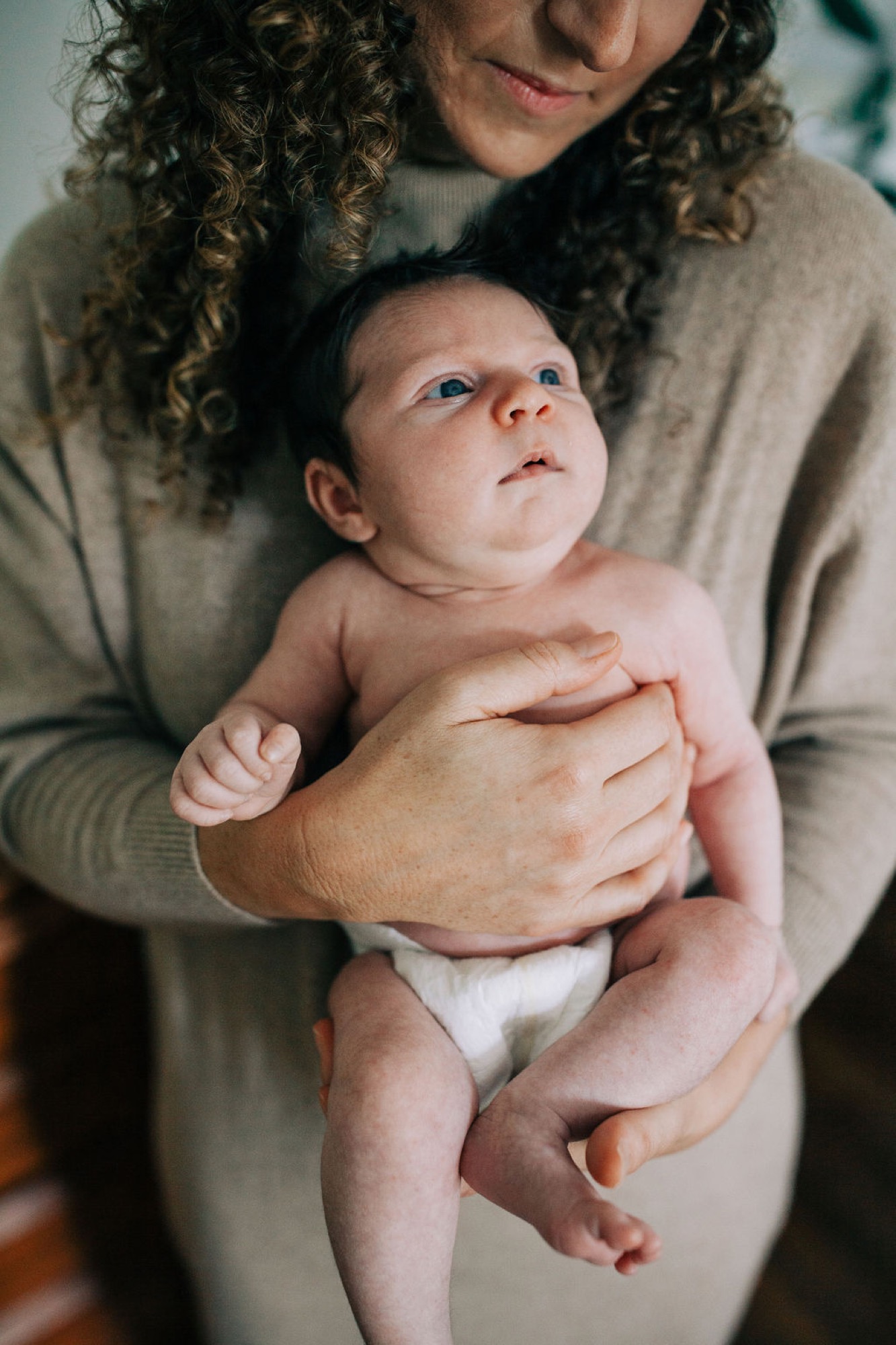 A newborn with bright blue eyes gazes upward while cradled in a parent's arms, soft natural light illuminating delicate features and tender hands