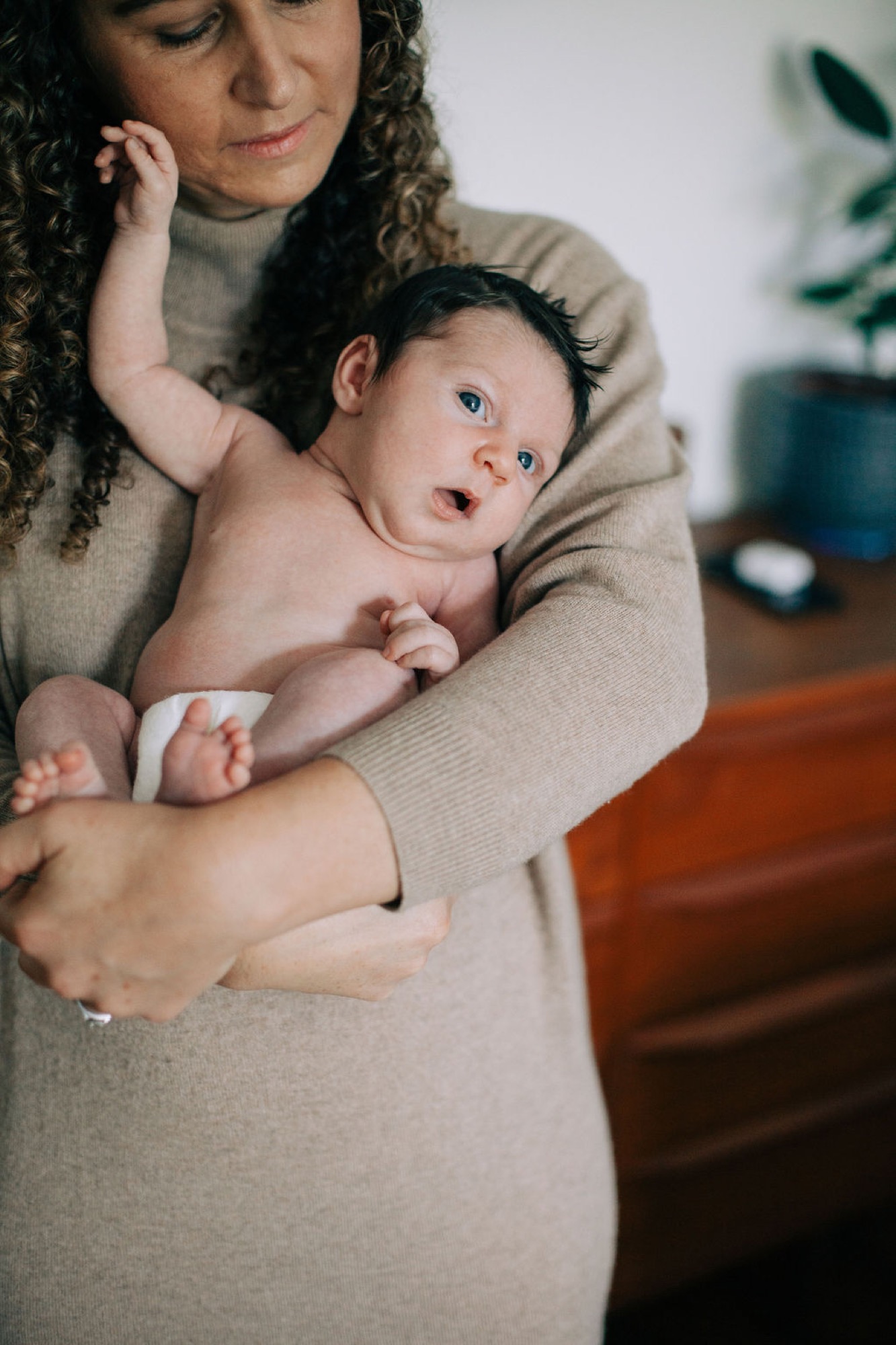 Mother in beige sweater gazes down at wide-eyed newborn she cradles, soft natural light illuminating their tender moment indoors