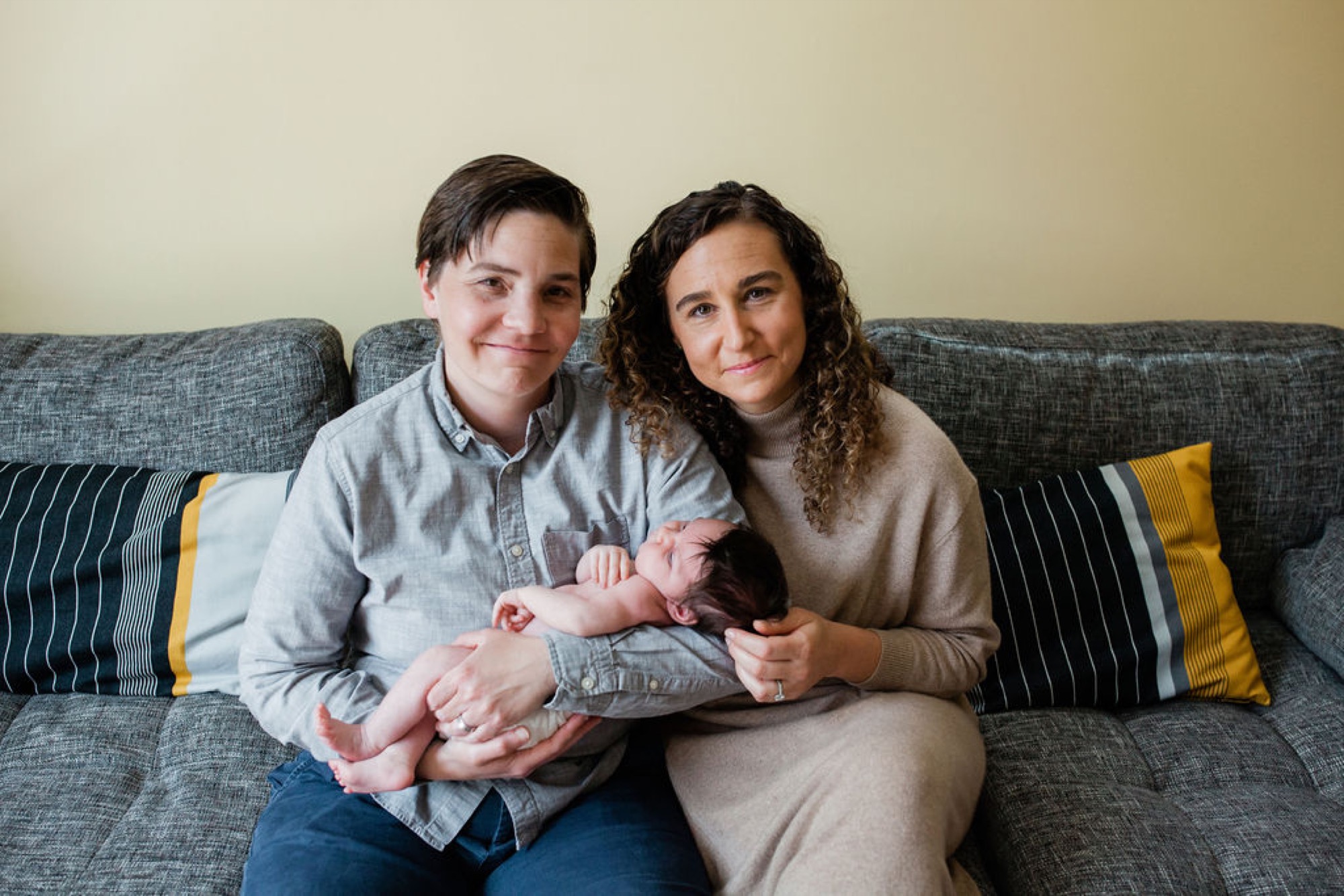 Two parents sit closely on a gray couch, gently cradling their newborn between them while smiling warmly at the camera