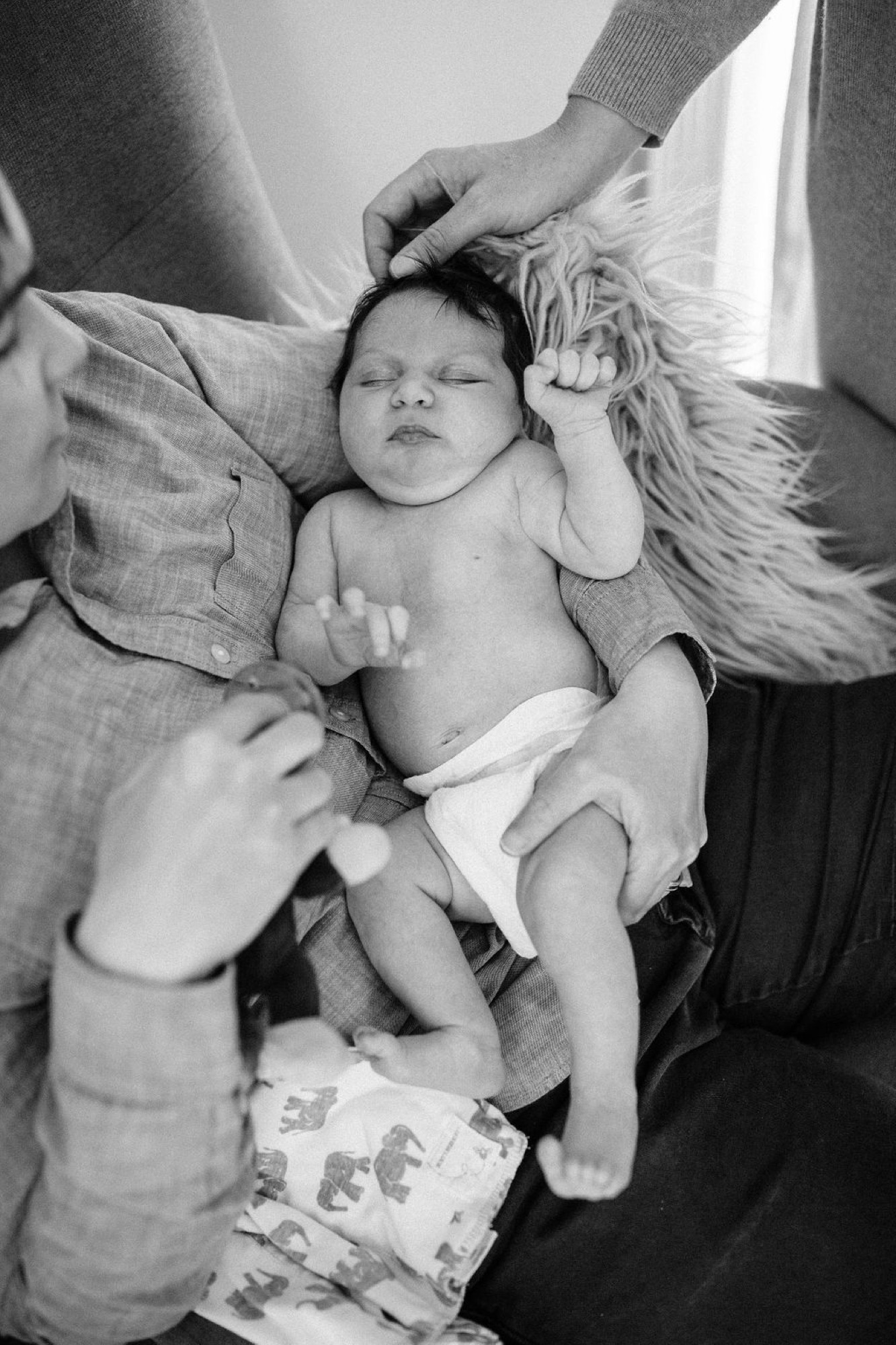 Sleeping newborn nestled between two adults in gray linen, one hand gently stroking the baby's head in intimate black and white portrait