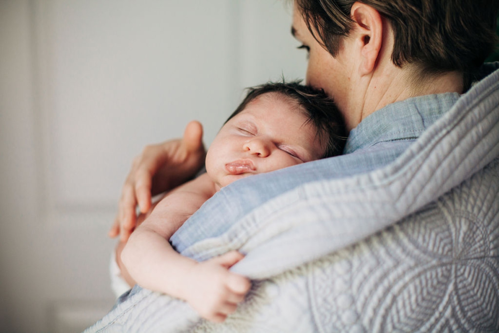 A parent in a blue striped shirt cradles their sleeping newborn against their chest, the infant's tiny hand resting peacefully on the caregiver's shoulder
