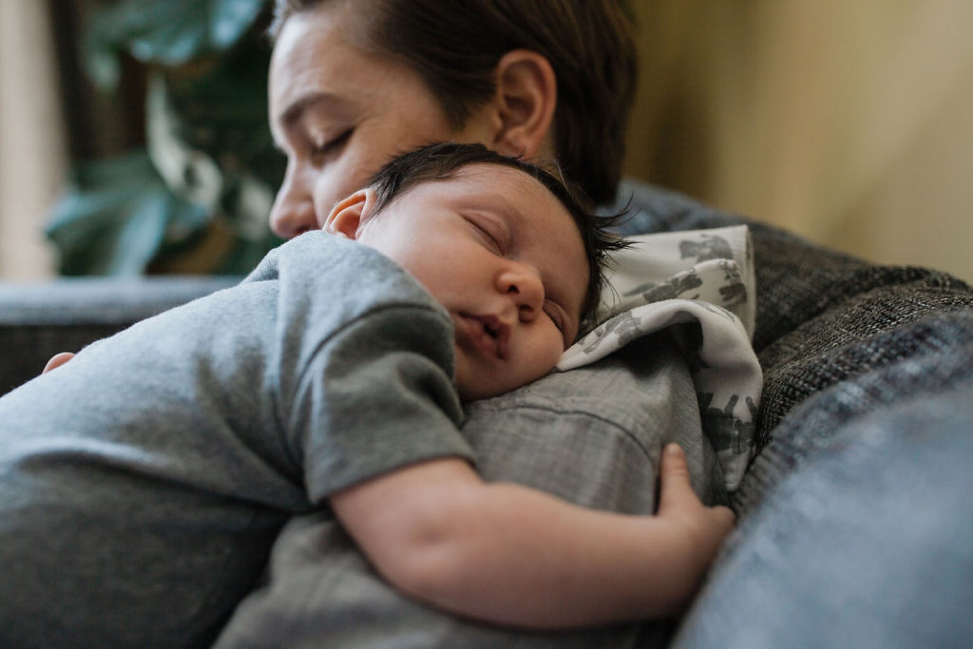 A mother cradles her sleeping newborn against her shoulder on a gray couch, both with eyes closed in serene contentment