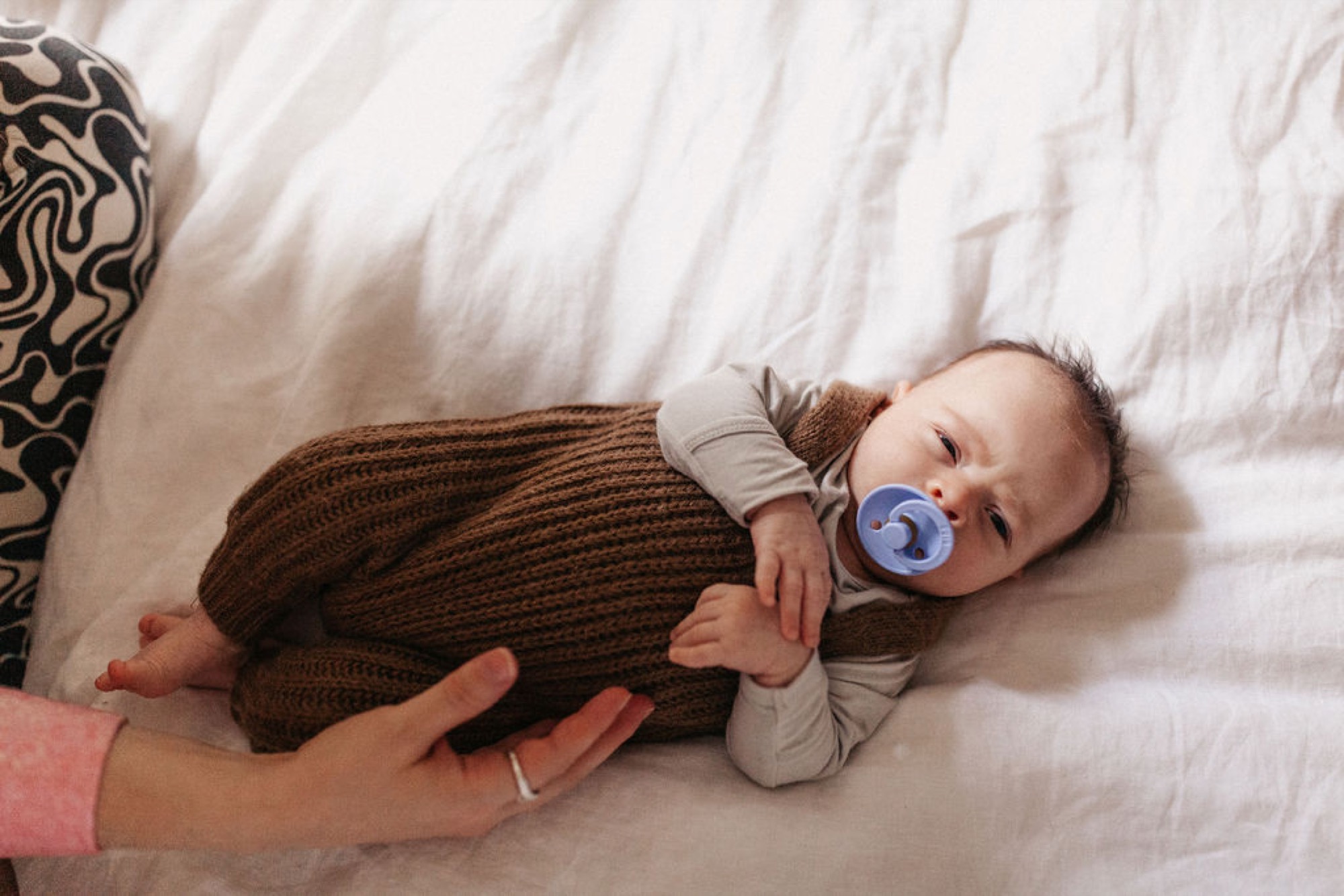 A newborn in a brown knit wrap rests on a white bedsheet while a parent's hand gently supports their body, pacifier in mouth