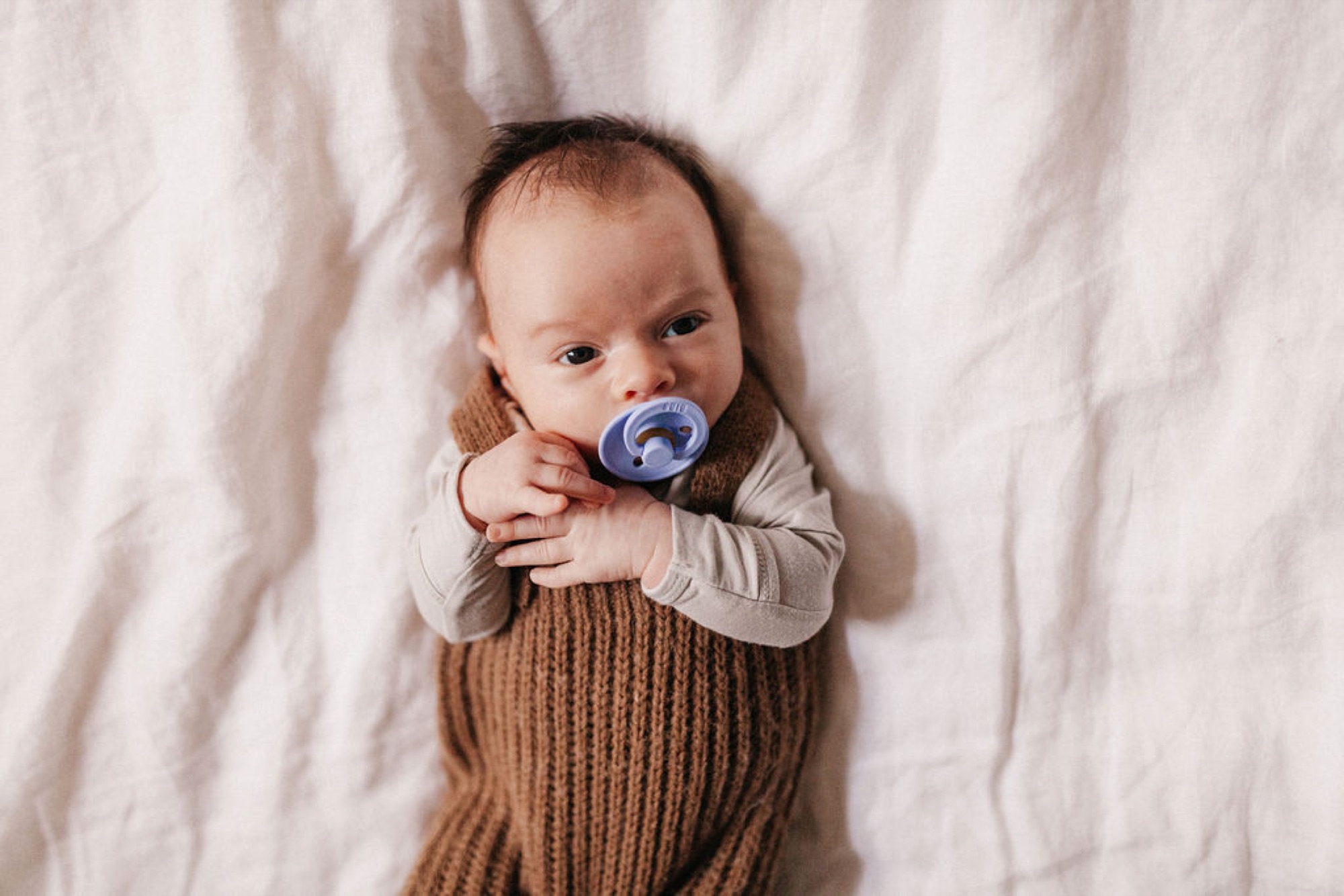 Newborn swaddled in a chunky knit wrap gazes upward while holding a blue pacifier against a cream linen backdrop