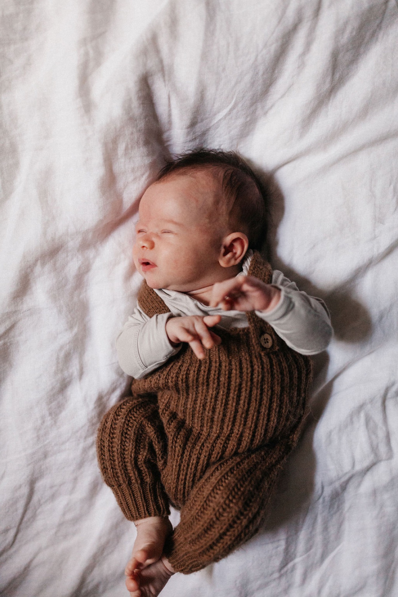 Newborn in brown ribbed knit pants sleeps peacefully on white linen, one arm raised with fingers splayed