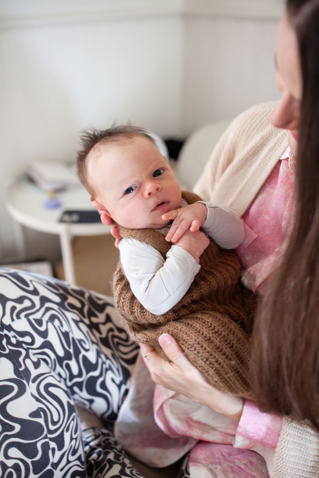 A newborn with alert blue eyes gazes upward while cradled in a parent's arms on a patterned bedspread