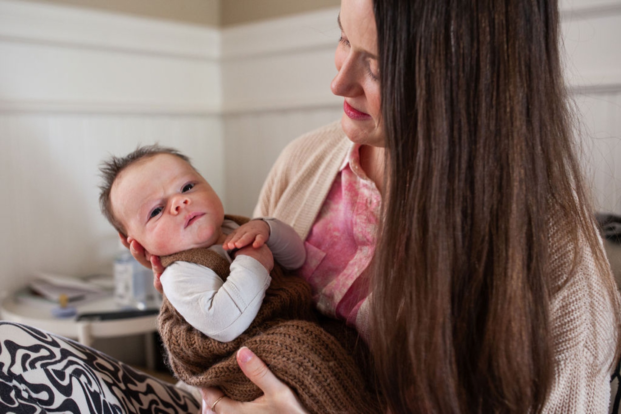 Mother gazes down at her newborn in a cream-toned nursery, soft natural light catching the baby's alert expression and delicate features
