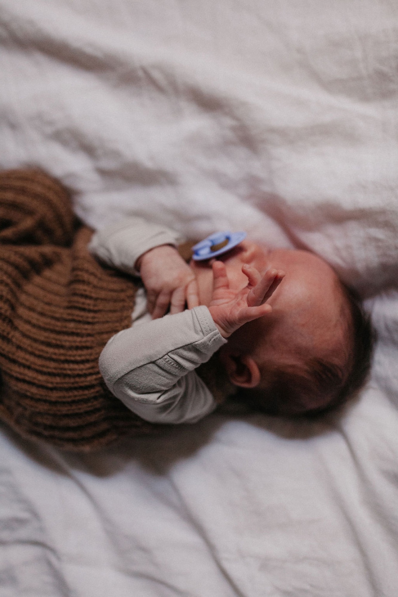 A newborn in a mustard knit sweater lies peacefully on white bedding, tiny hand grasping a blue pacifier against soft fabric