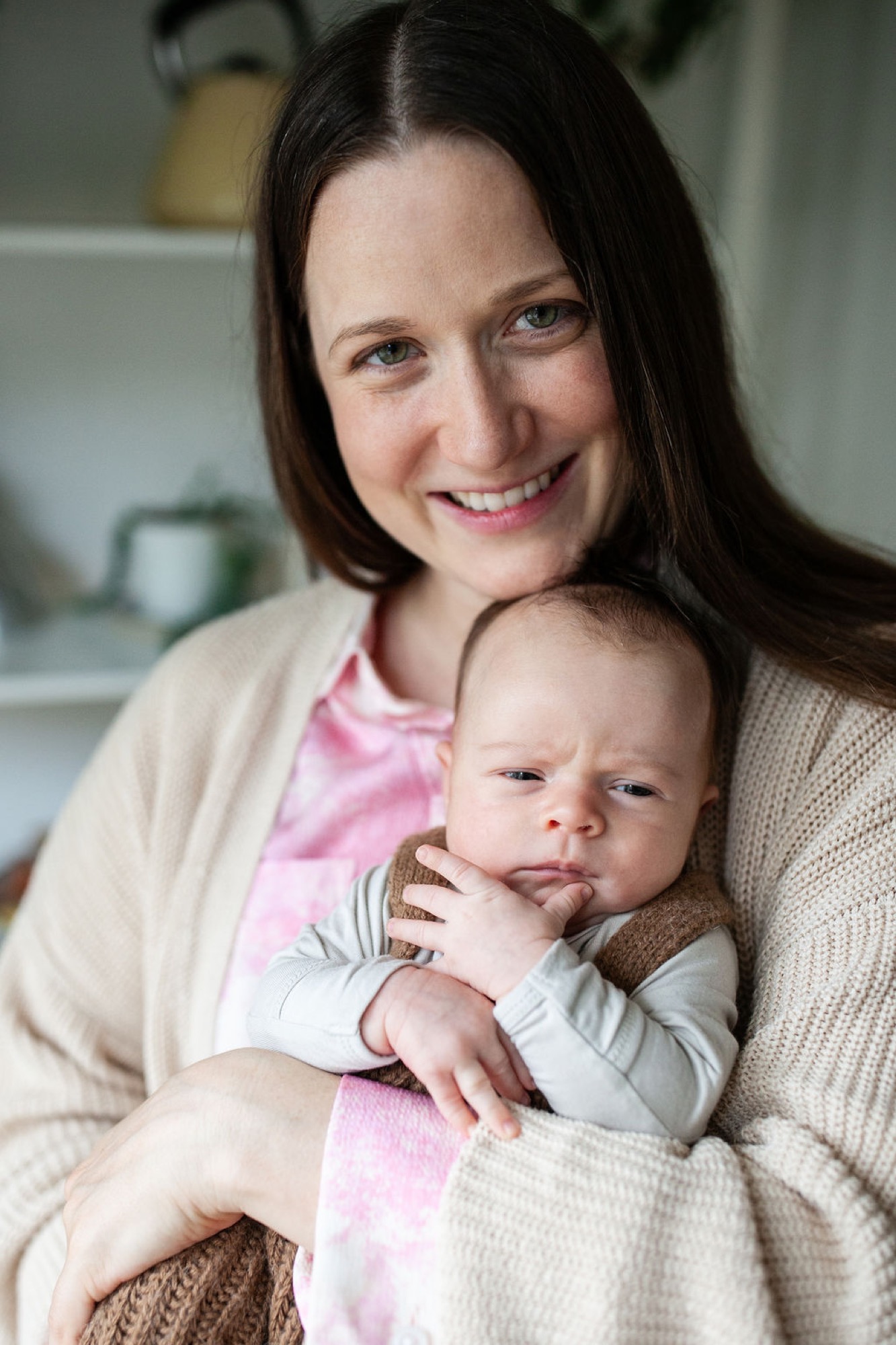 Mother in cream cardigan smiles warmly while cradling her sleeping newborn wrapped in striped clothing against soft natural indoor light