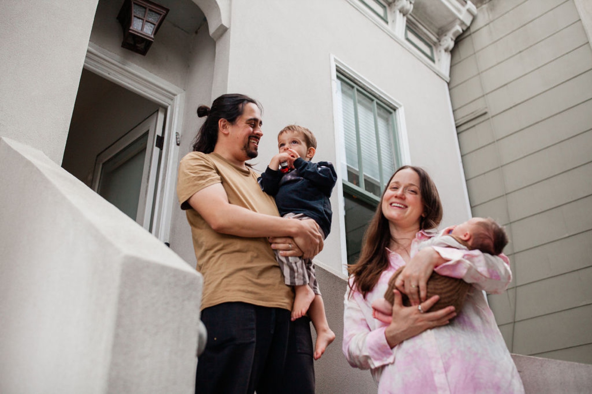 Two adults and two young children stand together in a bright white courtyard, smiling warmly while holding the babies close to their bodies