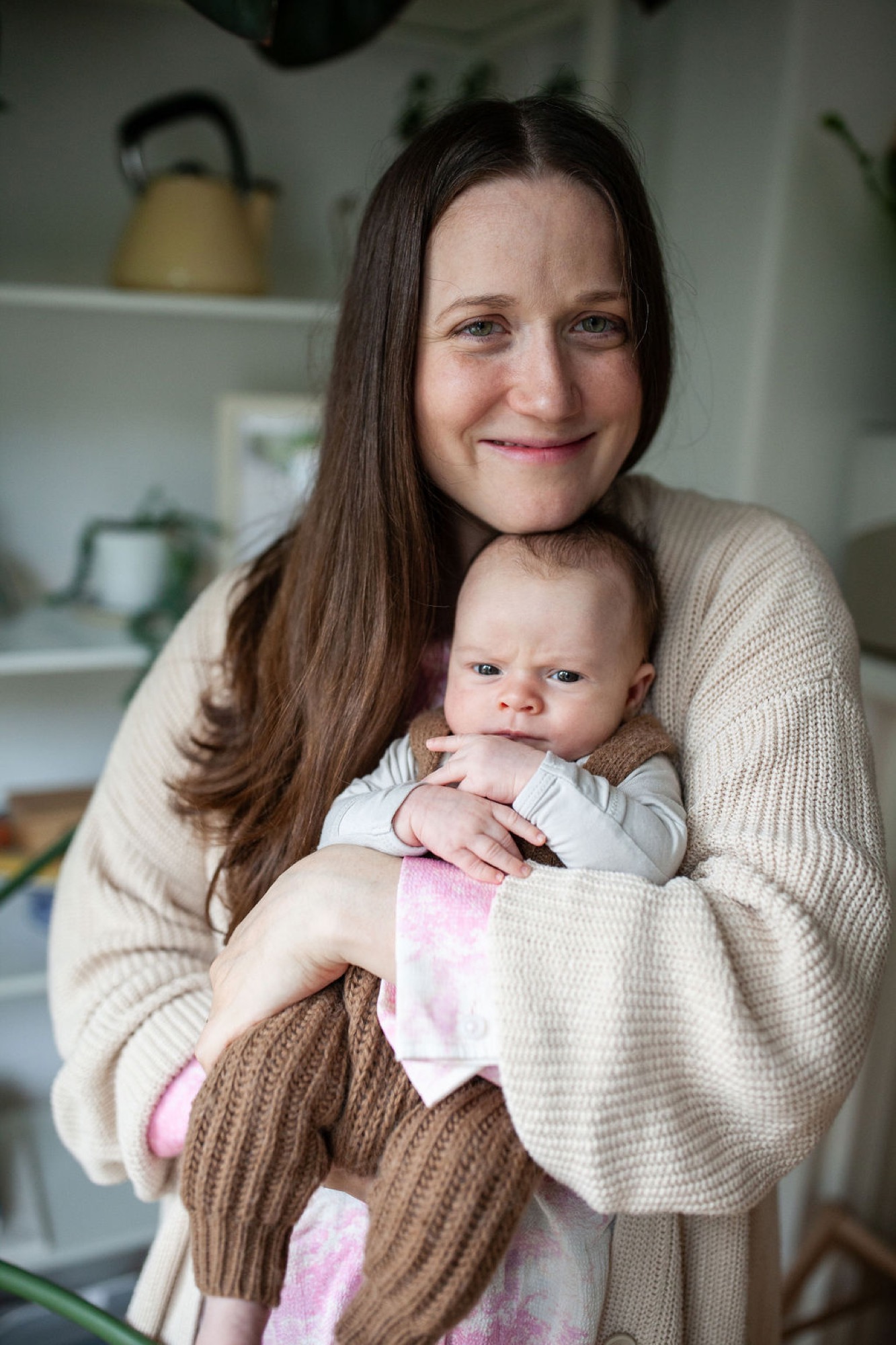 Mother in cream cardigan smiles warmly while cradling her newborn in a bright kitchen, soft natural light illuminating their gentle connection
