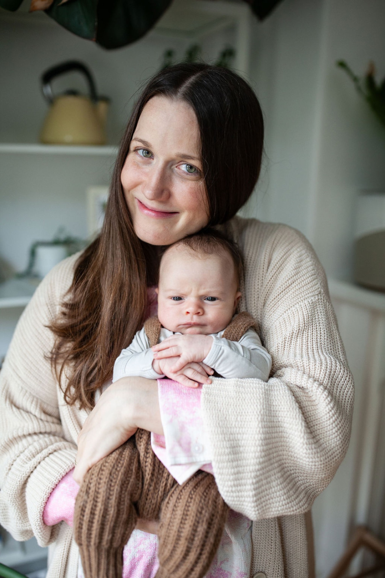 Mother in cream cardigan holds her infant with a gentle smile, soft natural light illuminating their faces in a bright kitchen with shelving behind them