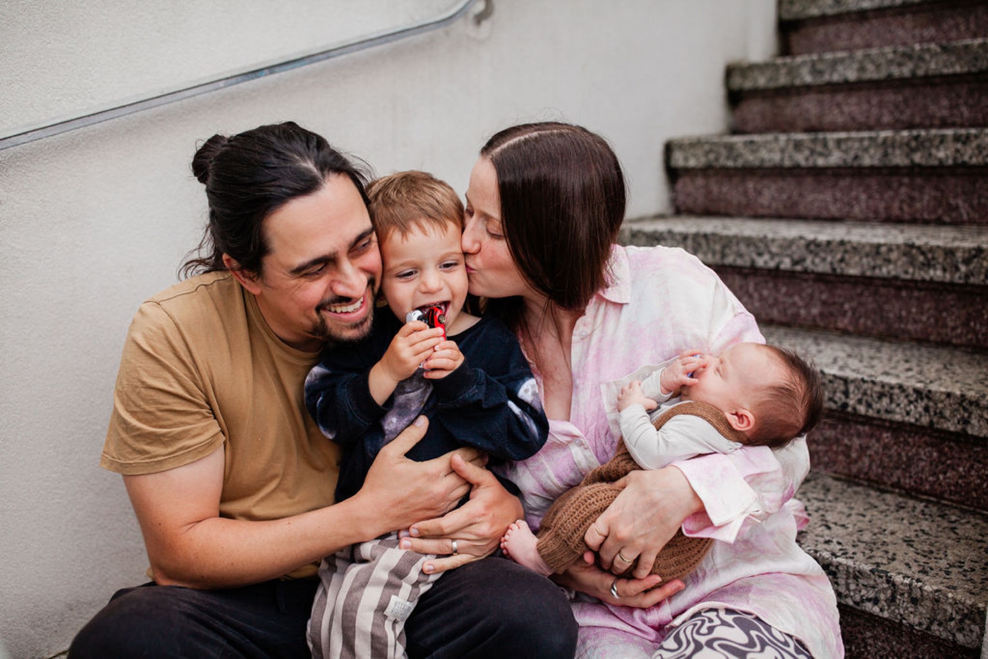A young family sits together on stairwell steps, the father holding their toddler while the mother kisses the newborn, all smiling warmly
