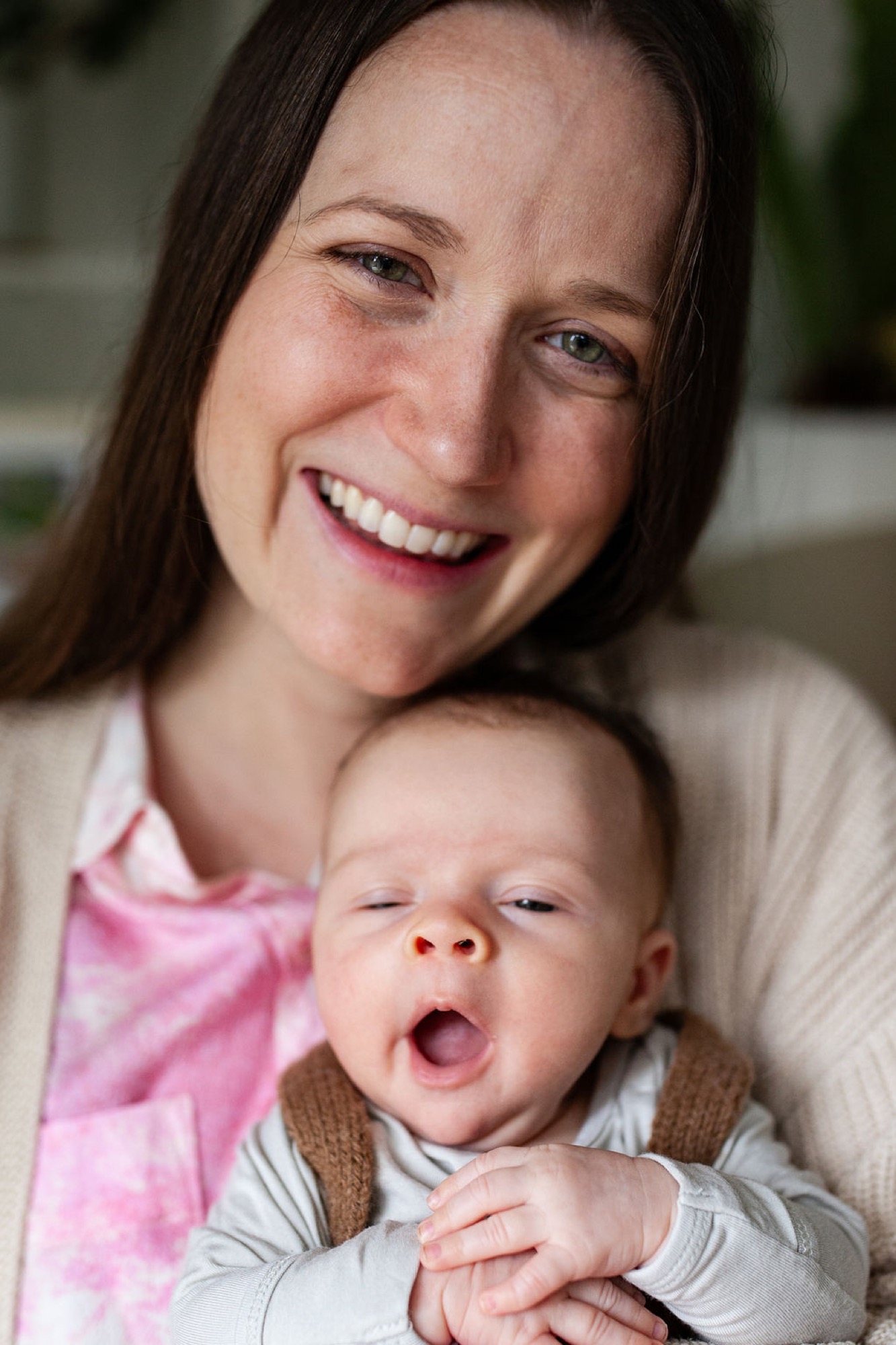 A smiling mother in pink holds her yawning newborn close, captured in warm indoor light during an intimate home session