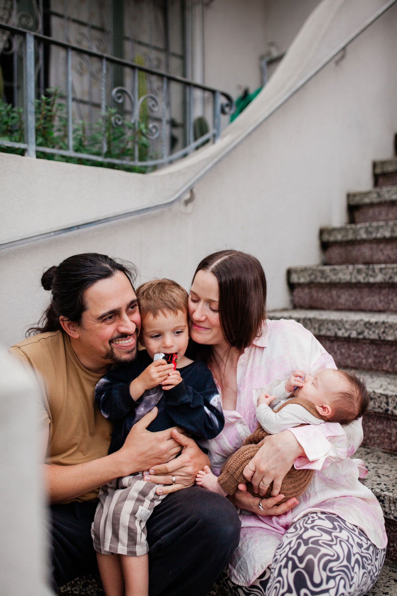 Family of four sits on curved stairs, parents smiling warmly while holding their toddler and newborn in soft natural light