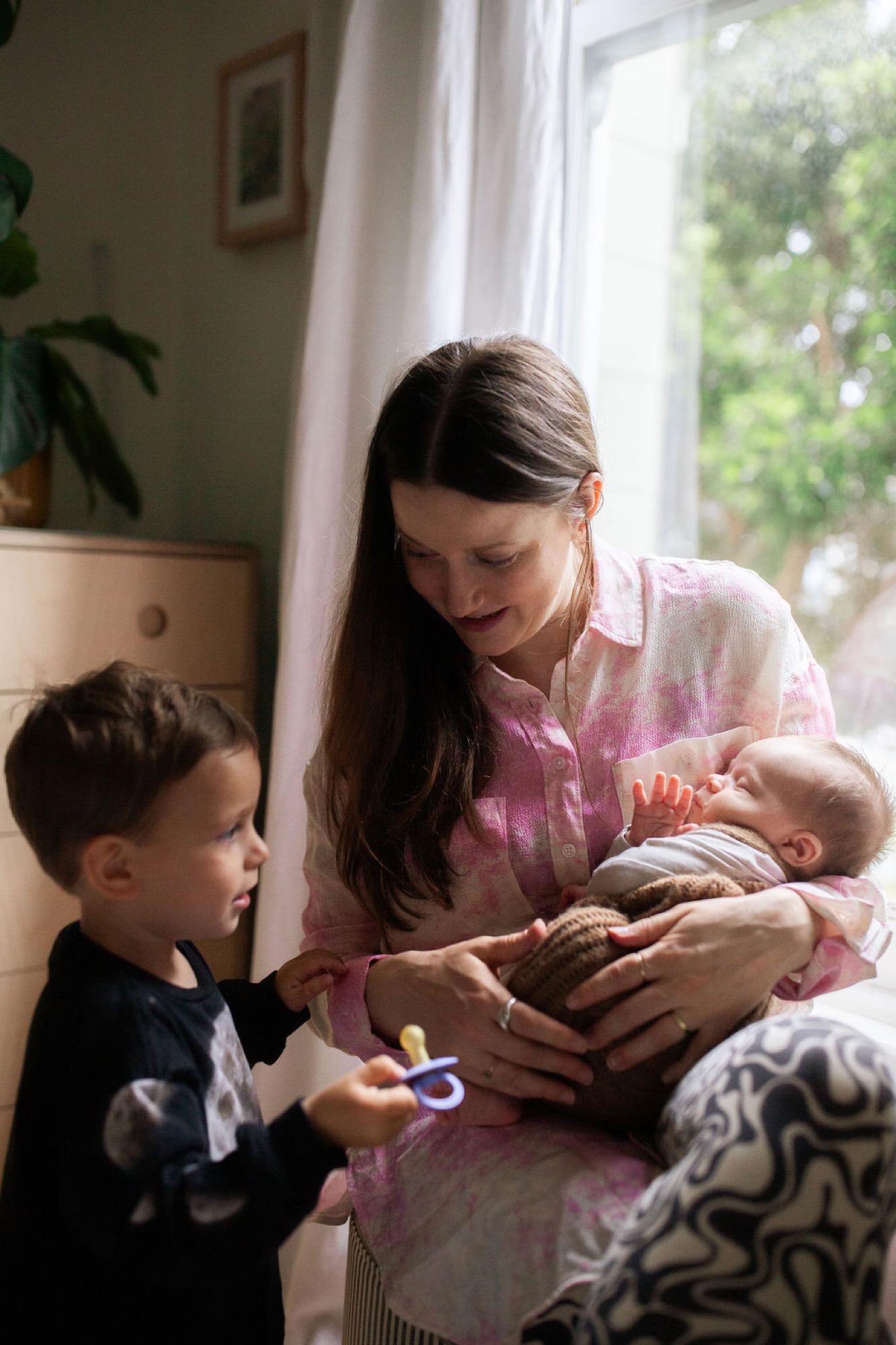 A mother in a pink tie-dye shirt holds her newborn while her toddler son in dark clothing reaches toward the baby by a sunlit window
