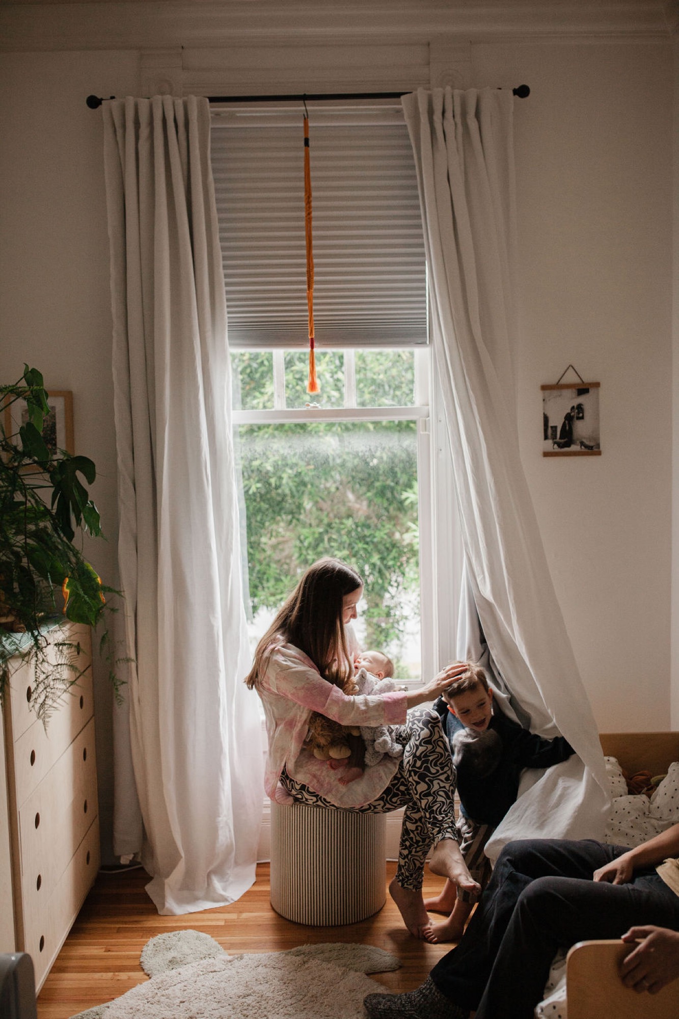 A woman in a rust-toned cardigan holds her infant while seated by a bright window with flowing white curtains, natural light illuminating their quiet moment together