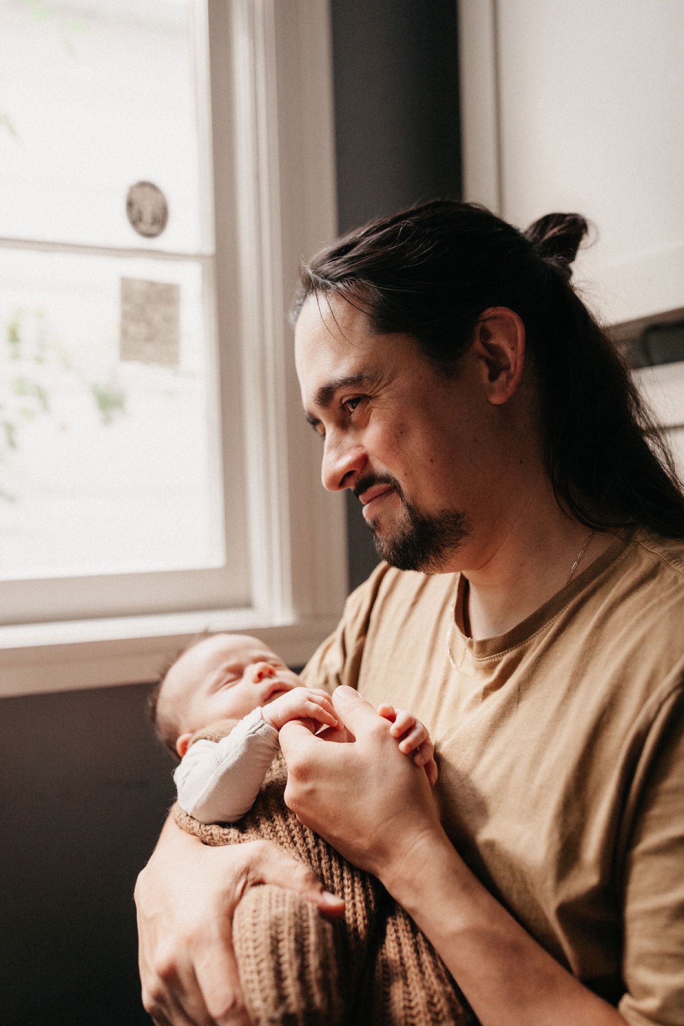 A father in a tan shirt gazes down at his newborn cradled in his arms by a sunlit window