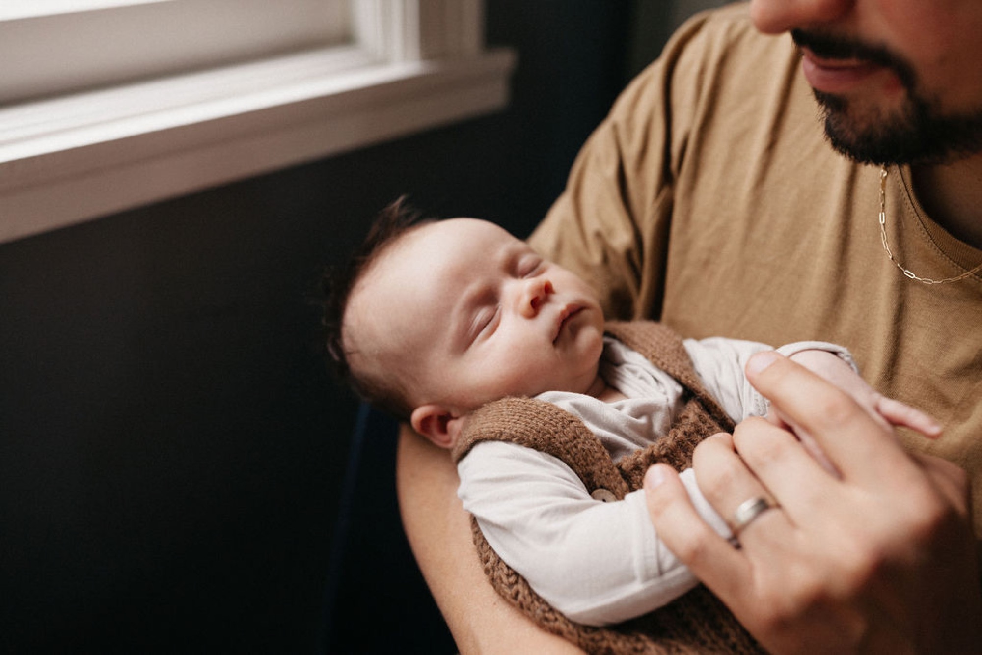 A father in a tan shirt gently cradles his newborn, who wears a brown knit wrap and gazes upward near a bright window