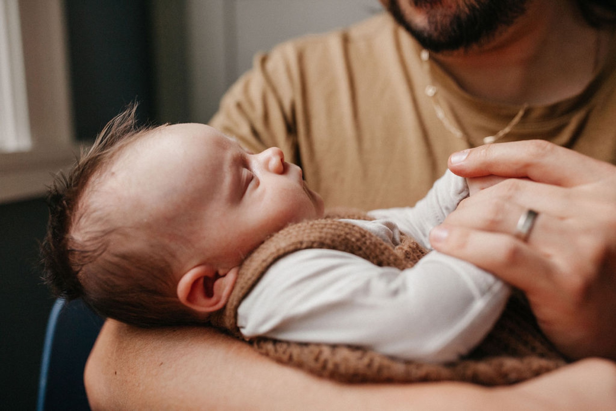 A bearded father in a tan shirt cradles his newborn close to his chest, the infant swaddled in a brown knit wrap against soft indoor light