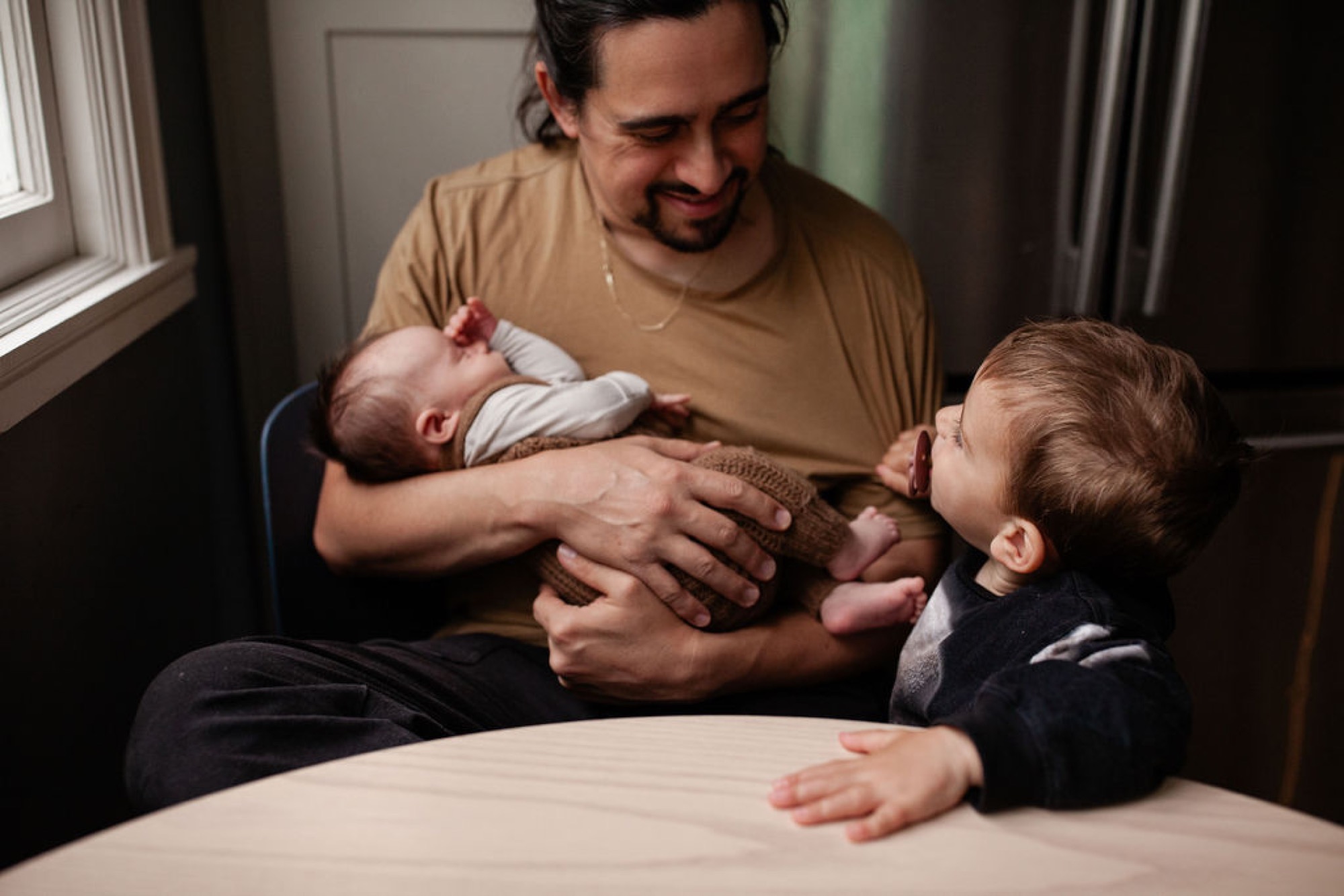 Father in tan shirt gently guides his toddler's hand toward newborn sibling wrapped in white, natural window light illuminating their tender interaction
