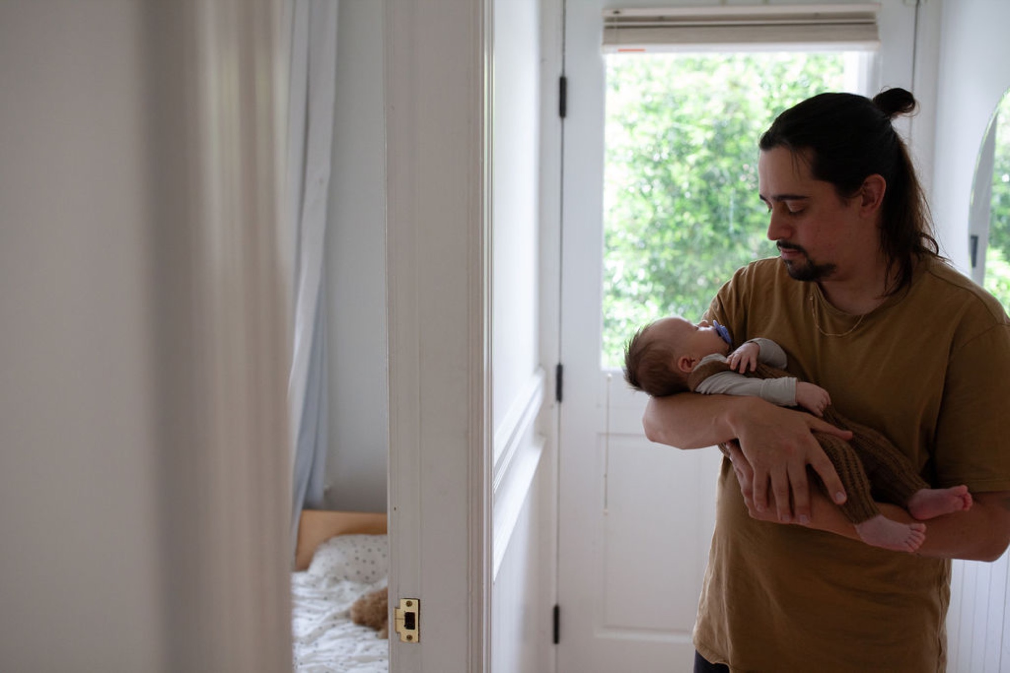Mother in golden shirt gazes down at newborn cradled in her arms by a bright doorway with sheer curtains