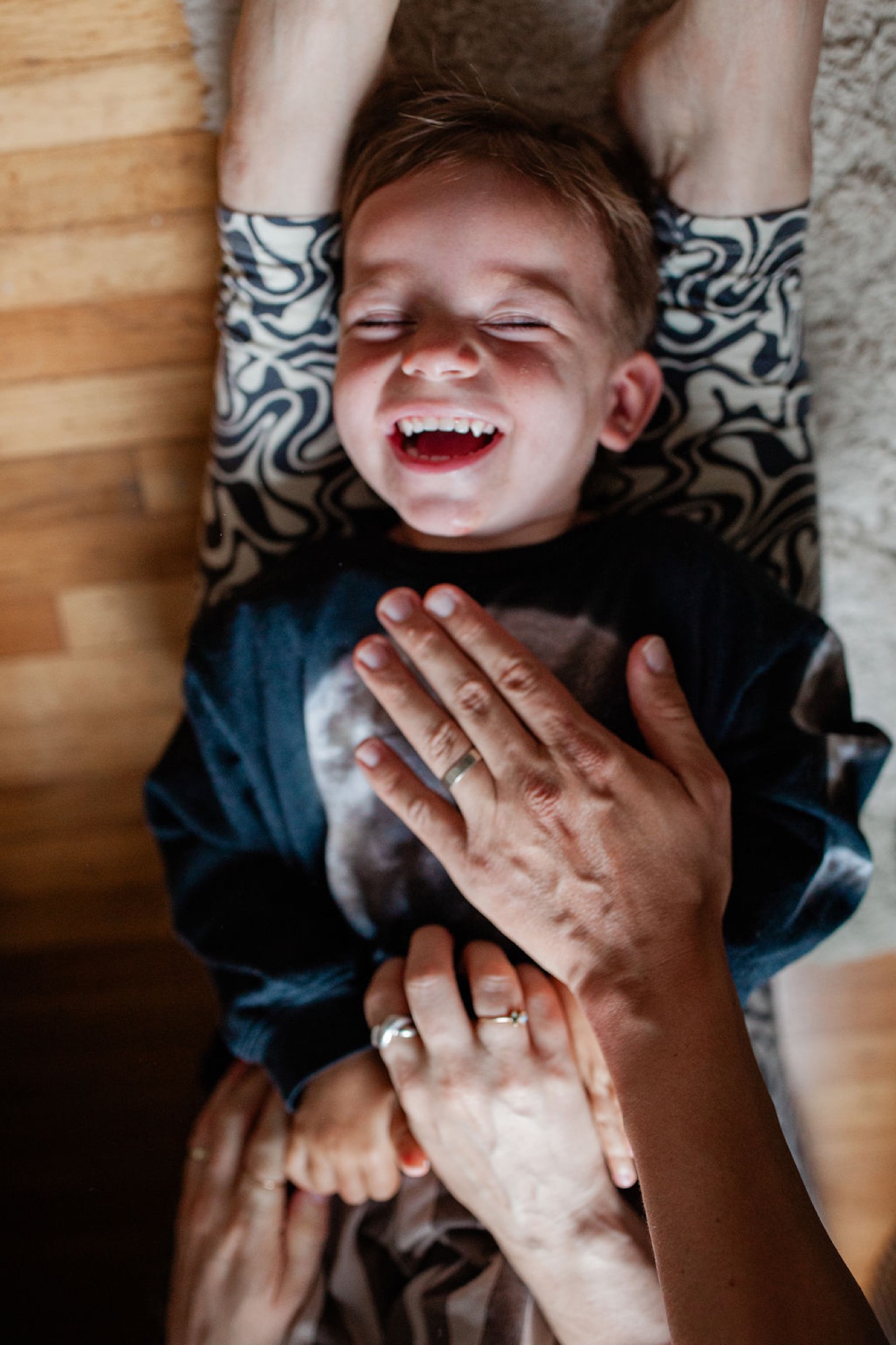 A laughing toddler lies on a patterned pillow while an adult's hands gently frame his joyful face from above