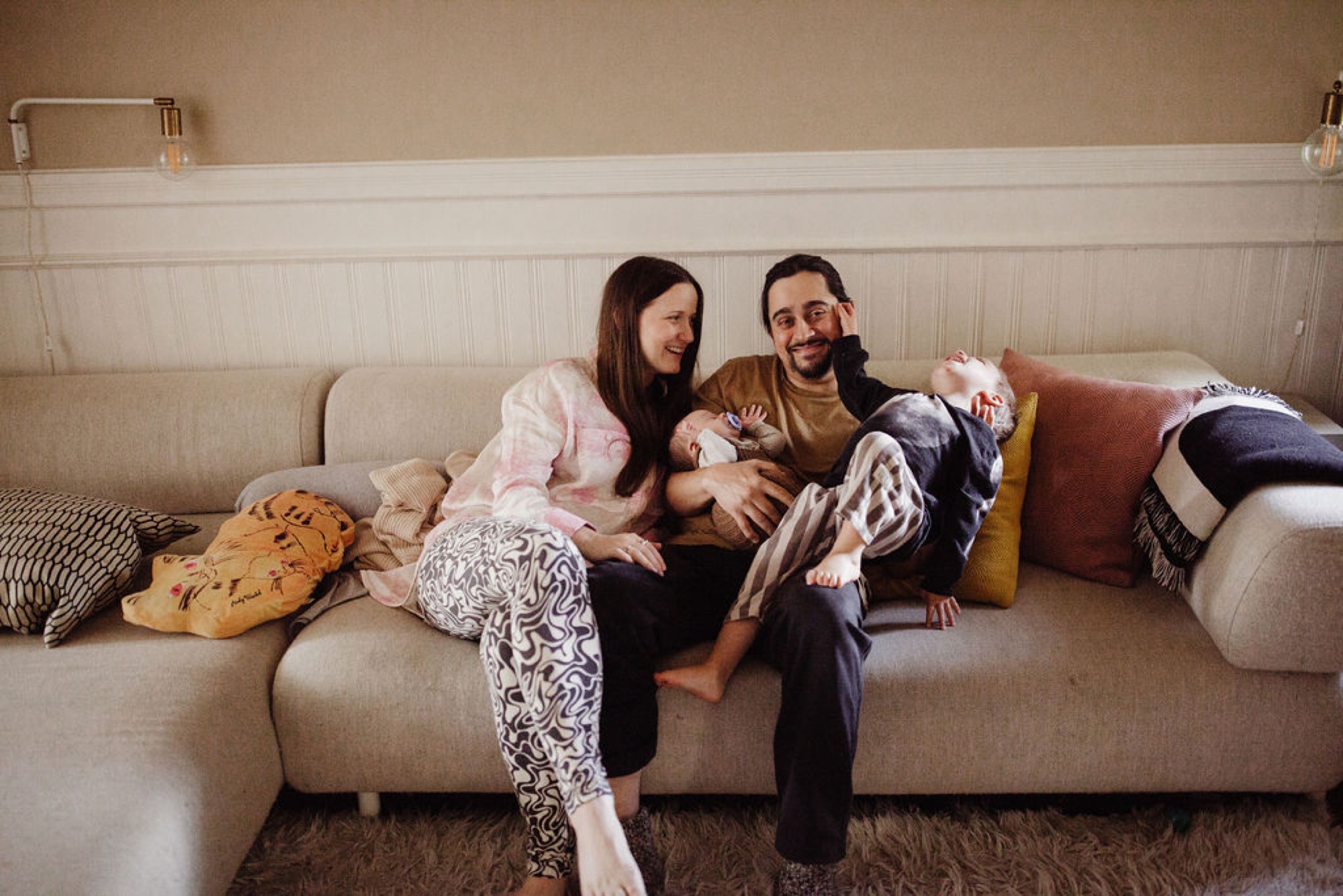 Parents sit together on a cream sofa surrounded by soft blankets, gazing down at their newborn with tender smiles in warm natural light