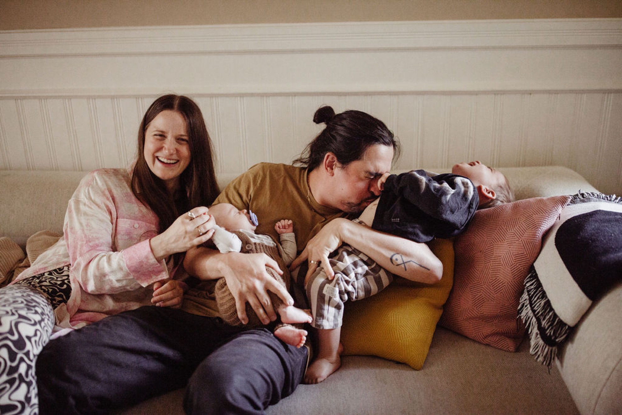 Two women sit on a cream couch with a newborn, their faces close and genuinely smiling while cradling the swaddled infant among colorful throw pillows